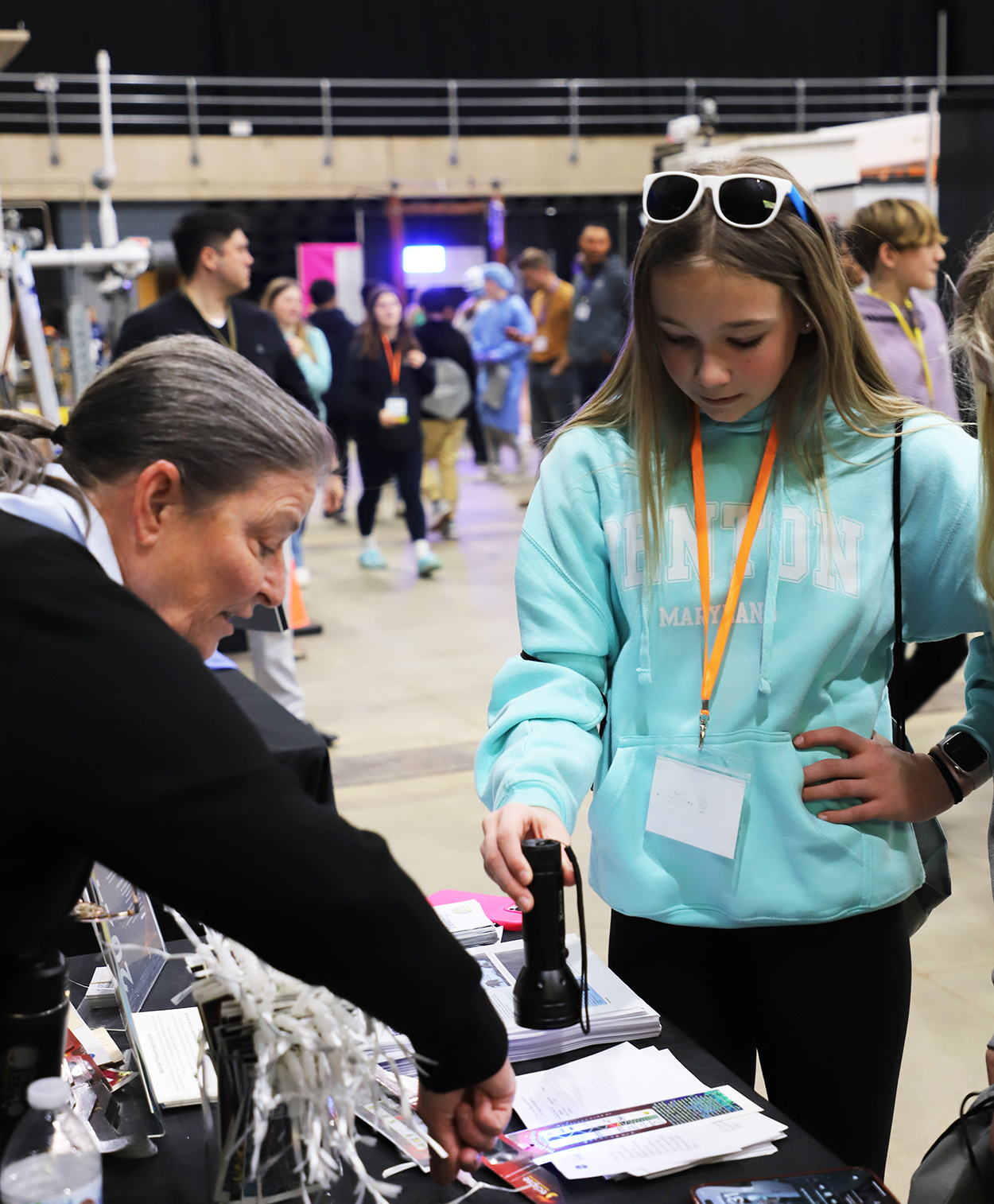 A person wearing a black shirt is showing a student in a teal sweatshirt how to hold a UV flashlight on beads inside a large center with exhibits and students walking in the background.