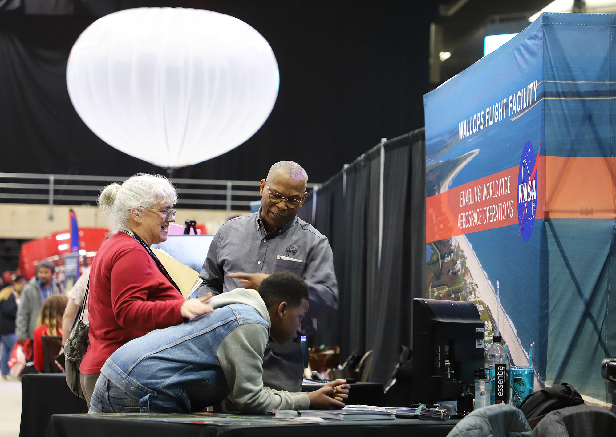 A person in a red shirt is smiling at a person from NASA and an eighth-grade child who is looking at a screen inside a center. A rocket range with ocean water and equipment is featured on a banner and a large white balloon is floating above.