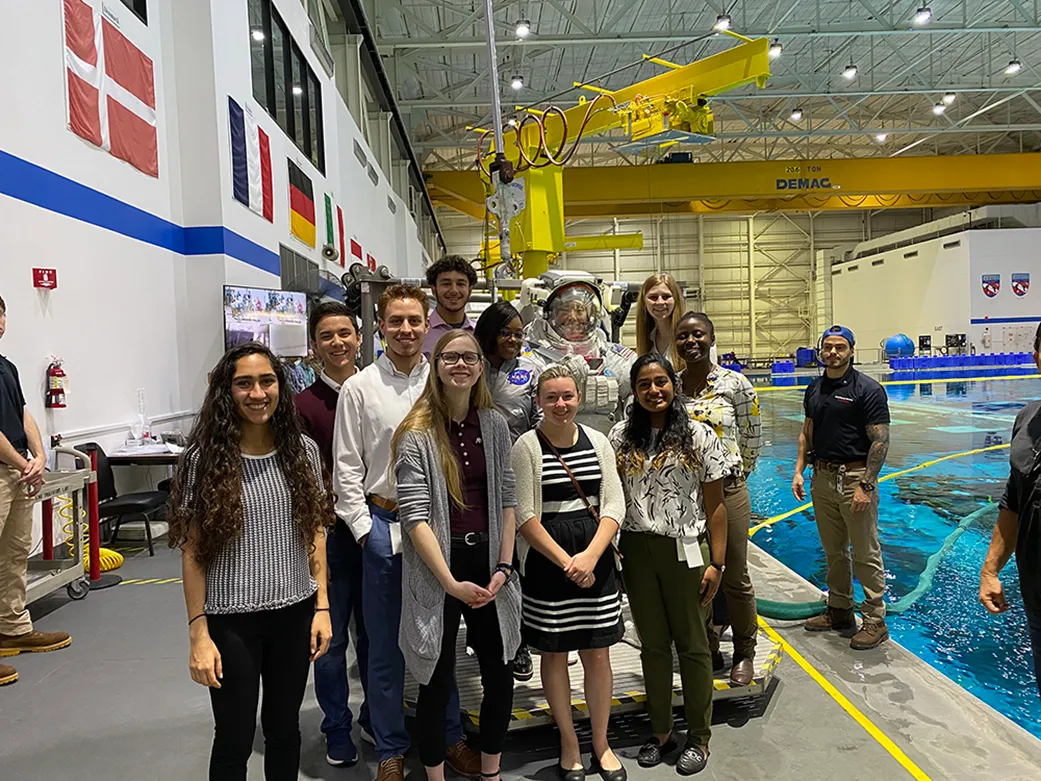 A group of students pose with astronaut Johnny Kim who is wearing a spacesuit. The group stands in front of a pool. 