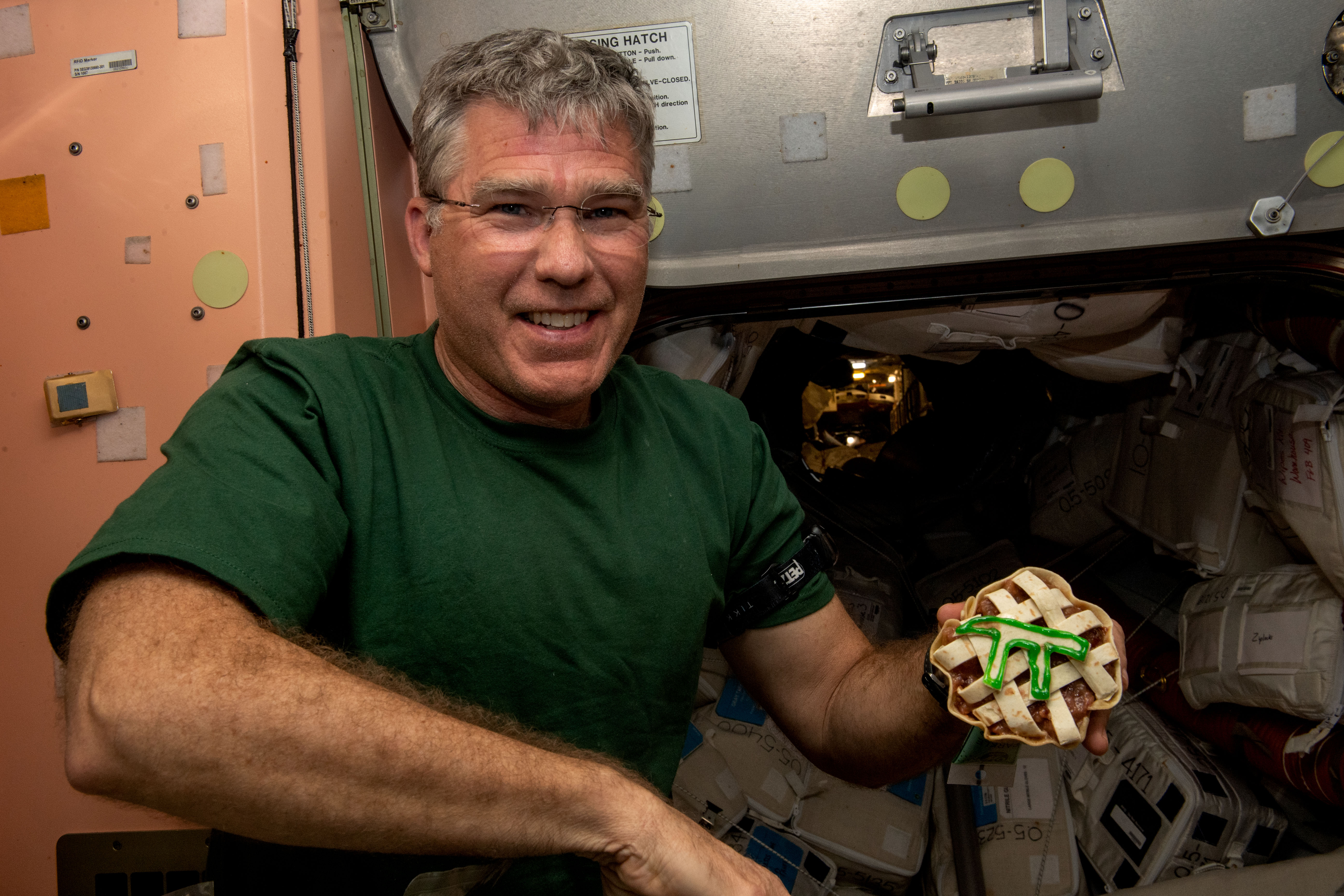 Stephen Bowen, wearing a green t-shirt and glasses, poses with a small latticed pie with a pi symbol outlined in green atop the lattice. Part of the International Space Station is visible behind him.