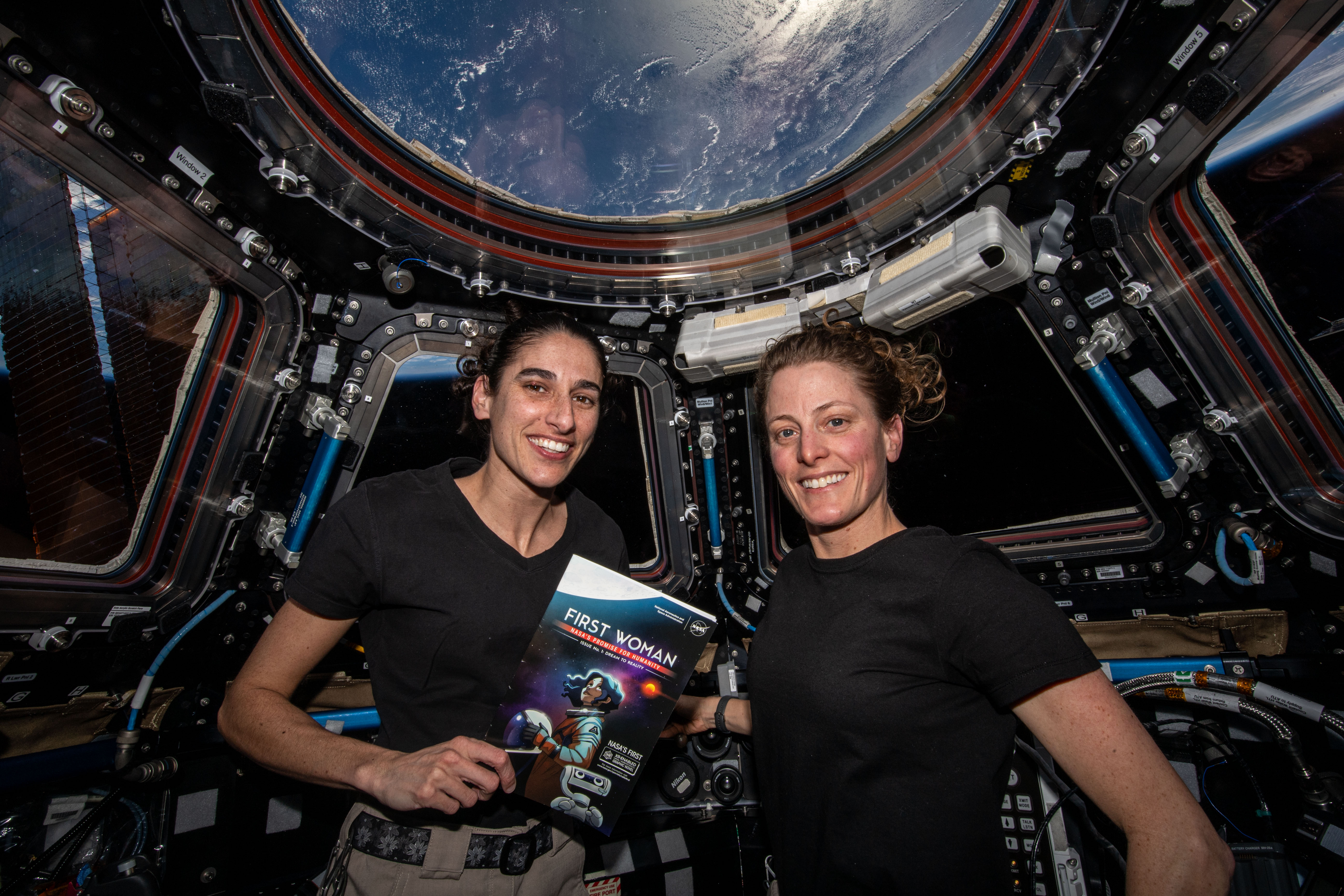 Two astronauts, Jasmin Moghbeli (left) and Loral O’Hara in the cupola of the International Space Station. The cupola has several windows; in the top round window, we can see Earth. The astronauts wear black t-shirts and smile at the camera. Moghbeli holds a copy of First Woman, NASA's official graphic novel about a fictional astronaut who is the first woman to explore the Moon.
