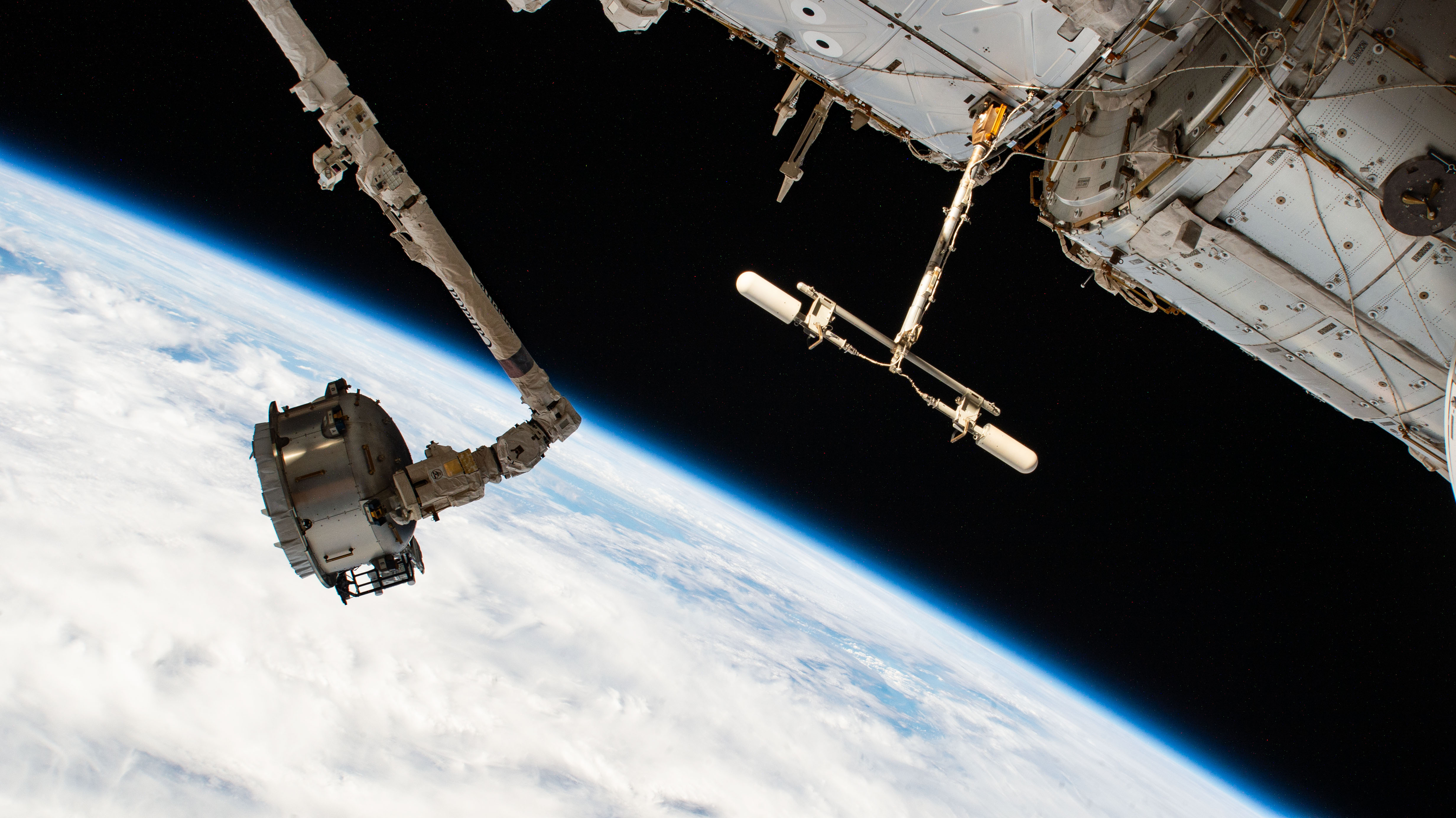 At left, the Canadarm2 robotic arm maneuvers the NanoRacks Bishop airlock in the grip of its latching end effector. At top right, a UHF antenna extends from the forward end of the Destiny laboratory module, which is also attached to the Harmony module.