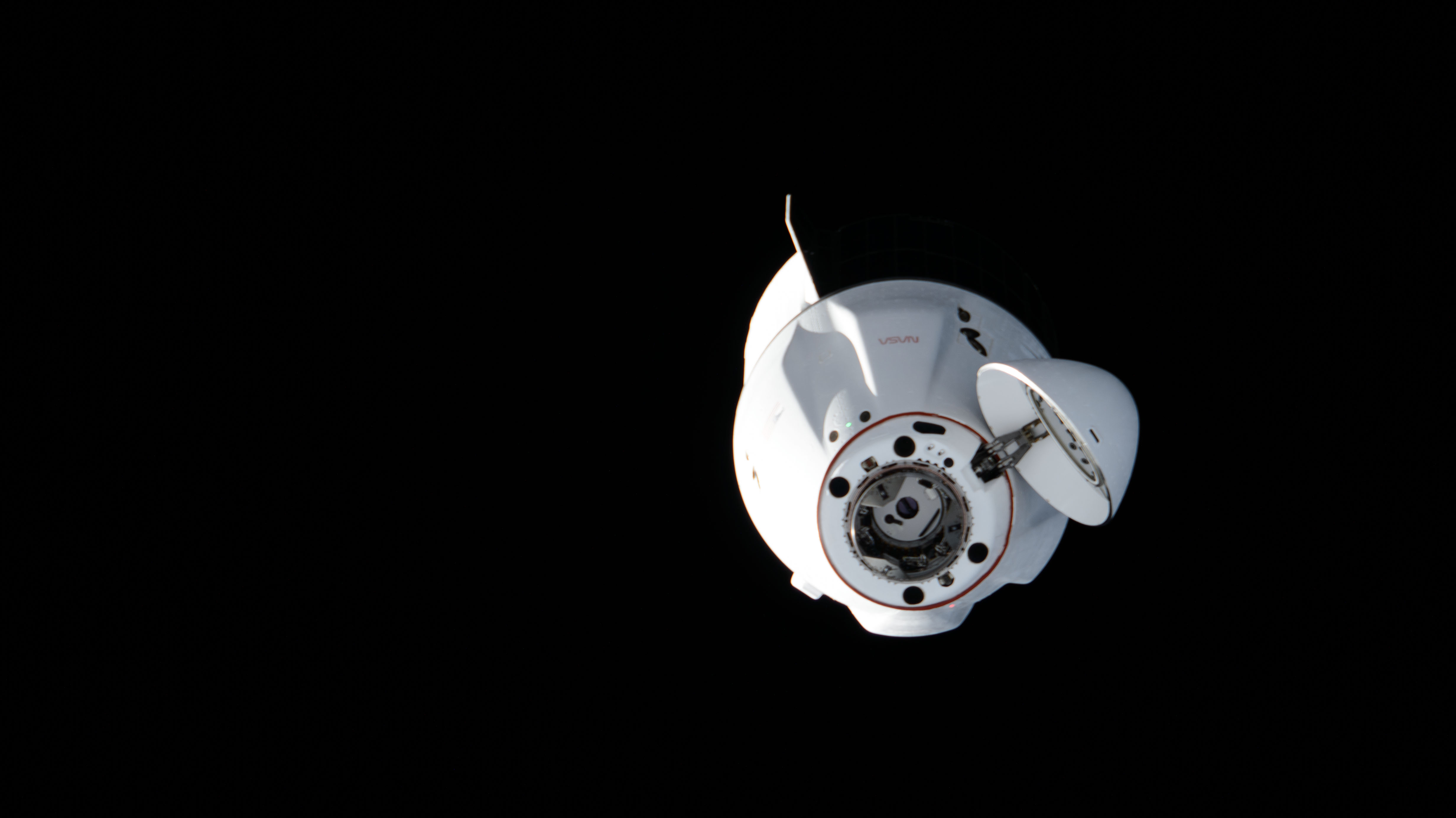 The SpaceX Dragon cargo craft approaches the International Space Station for a docking to the Harmony module's space-facing port. The spacecraft is a stark white against the black background of space, with its nosecone open and ready to dock.