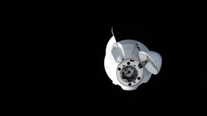 The SpaceX Dragon cargo craft approaches the International Space Station for a docking to the Harmony module's space-facing port. The spacecraft is a stark white against the black background of space, with its nosecone open and ready to dock.