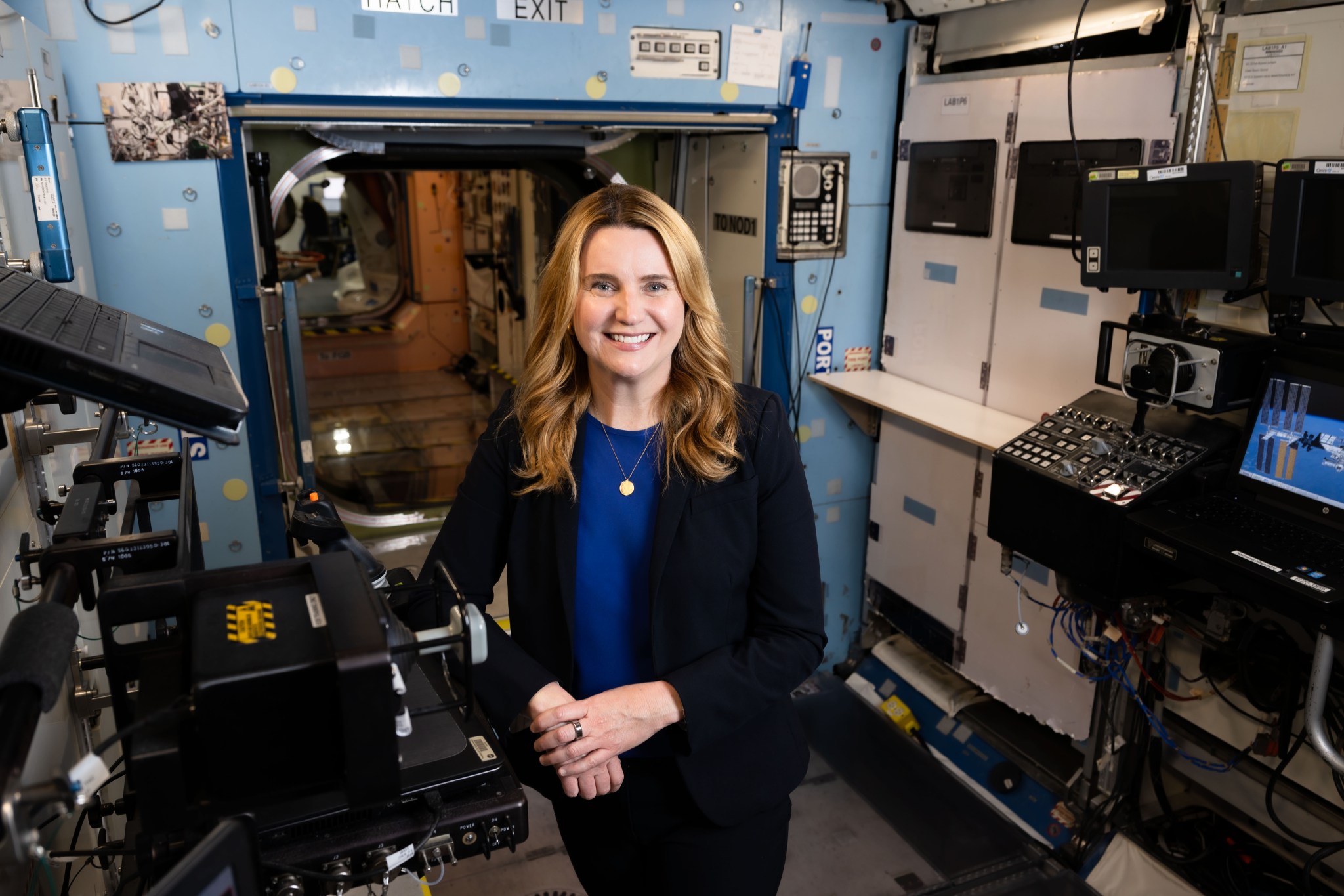 A woman with wavy blonde hair slightly past her shoulders smiles at the camera with one hand over the other as she leans against equipment inside one of the International Space Station modules inside the Space Vehicle Mockup Facility at NASA's Johnson Space Center. She is wearing black dress pants, a blue blouse, and a black jacket, and a circular pendant is visible around her neck. Black computer equipment and white cabinets are seen against the left and right walls of the module, and a large rounded square opening to other modules is present behind Jennifer. The wall surrounding the opening is light blue, and there are many items and labels affixed to it. Blue handrails are present on both sides of the opening, as well as on one of the walls.