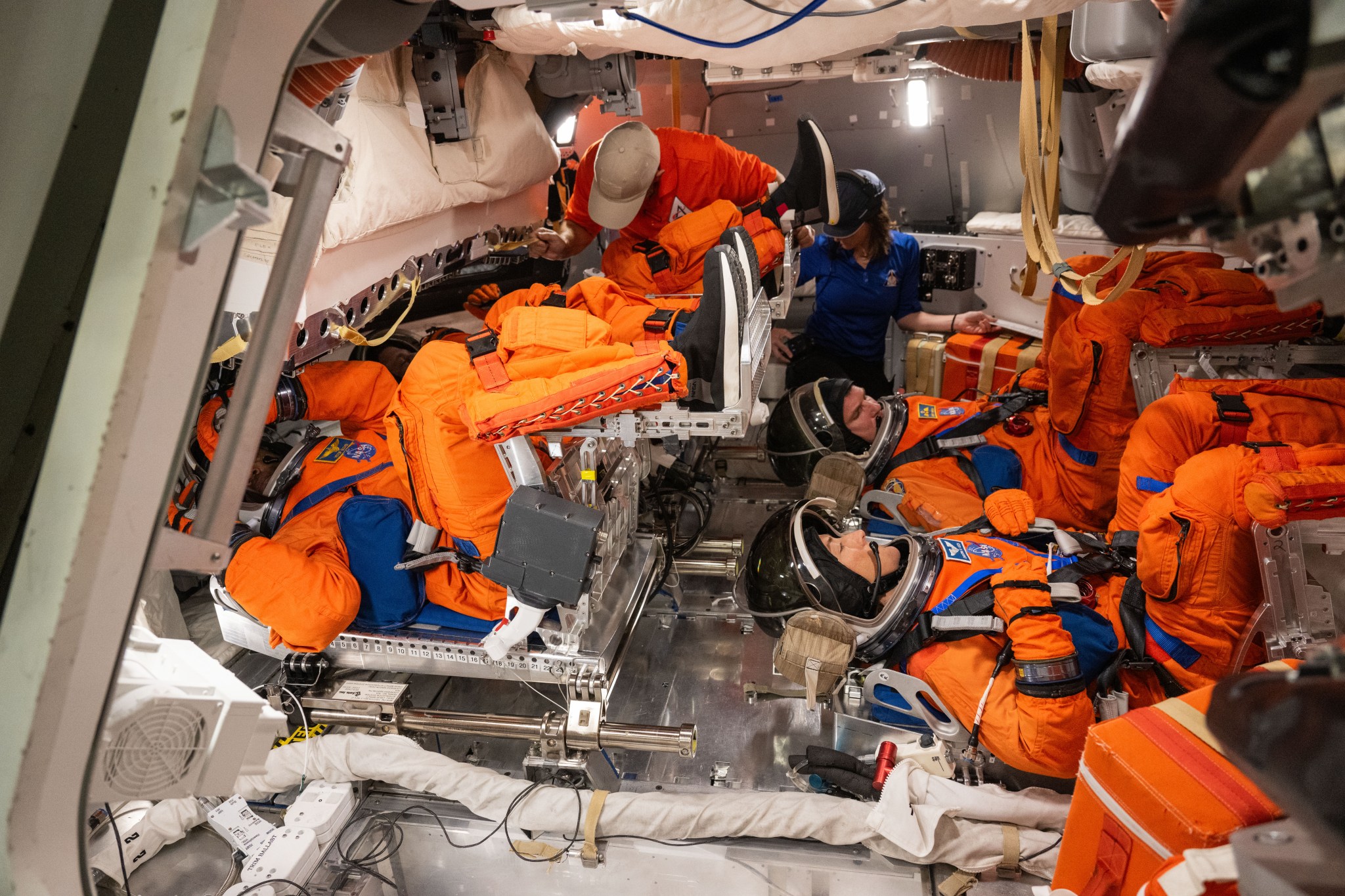 Four people dressed in orange spacesuits sit in a mockup of a spacecraft.