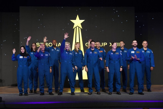 The latest astronaut candidate graduates, a group of men and women of different races and ethnicities, greet the audience (not pictured). The candidates all wear blue jumpsuits with patches on them. Behind them is a black and gold graphic of a star streaking upwards. The background has white dots on it that resemble distant stars.
