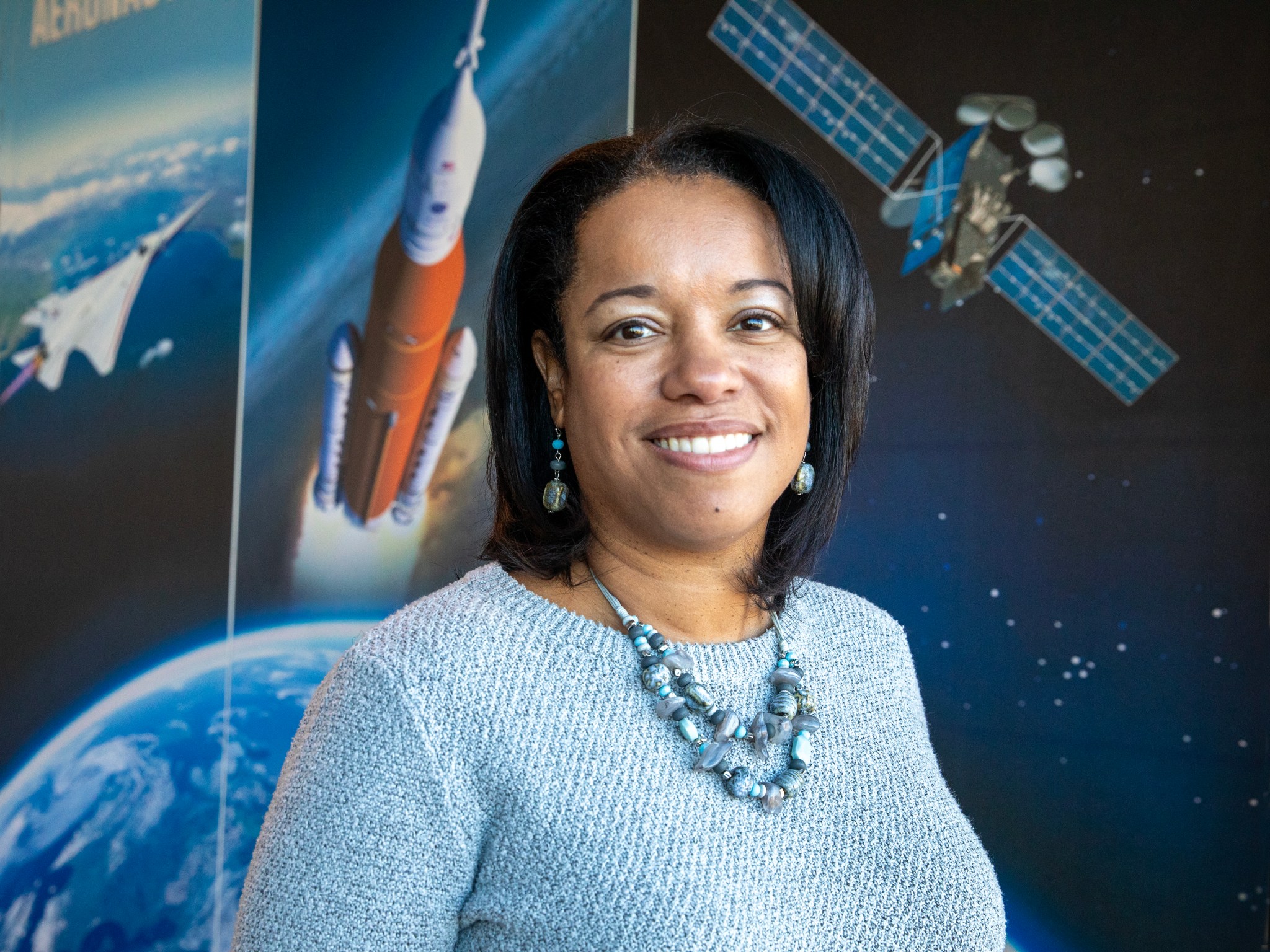 Dr. Kanama Bivins is Acting Associate Director at NASA Langley Research Center. She is pictured here smiling and wearing a grey sweater and necklace. She is standing in front of a poster featuring imagery from NASA missions, including Artemis and