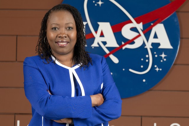 A woman with twisted dark brown hair and brown eyes smiles happily at the camera while crossing her hands across her chest. She's wearing a royal blue blazer with white piping around the outside. She stands before a brick wall with the NASA logo behind her.