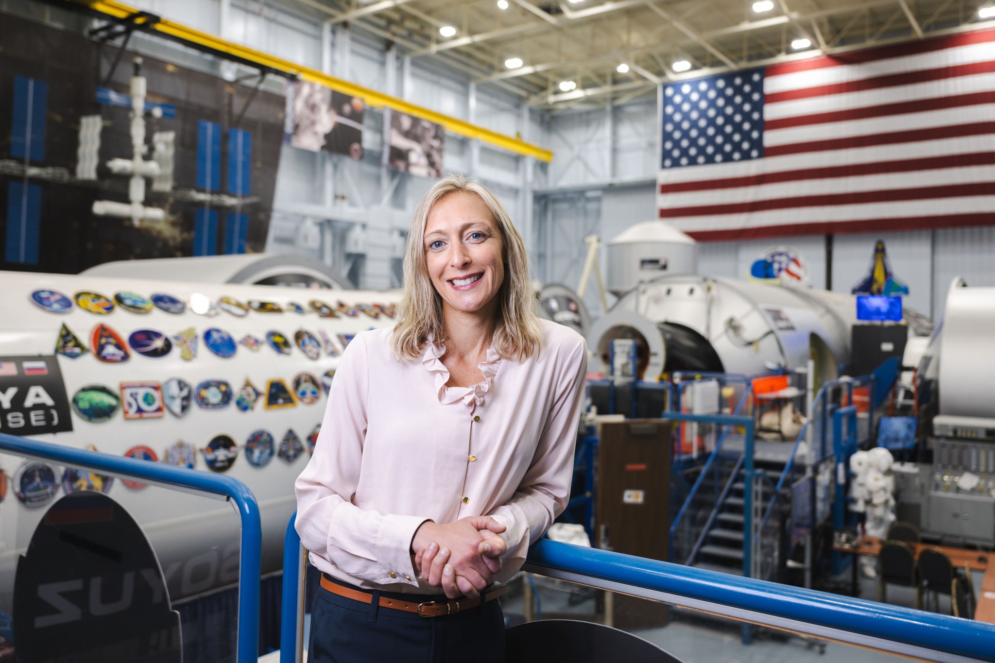 A woman with straight, shoulder-length blonde hair smiles at the camera with her hands clasped together as she leans on a blue safety railing overlooking the International Space Station modules inside the Space Vehicle Mockup Facility at NASA's Johnson Space Center. She is wearing dark dress pants, a brown belt, and a light-colored blouse with buttons and a ruffled collar. Many flight patches are visible on the white exterior of the cylindrical modules visible below her. A large American flag hangs from the ceiling in the top right of the image, with the patches of STS-51-L and STS-107 on the wall beneath it.