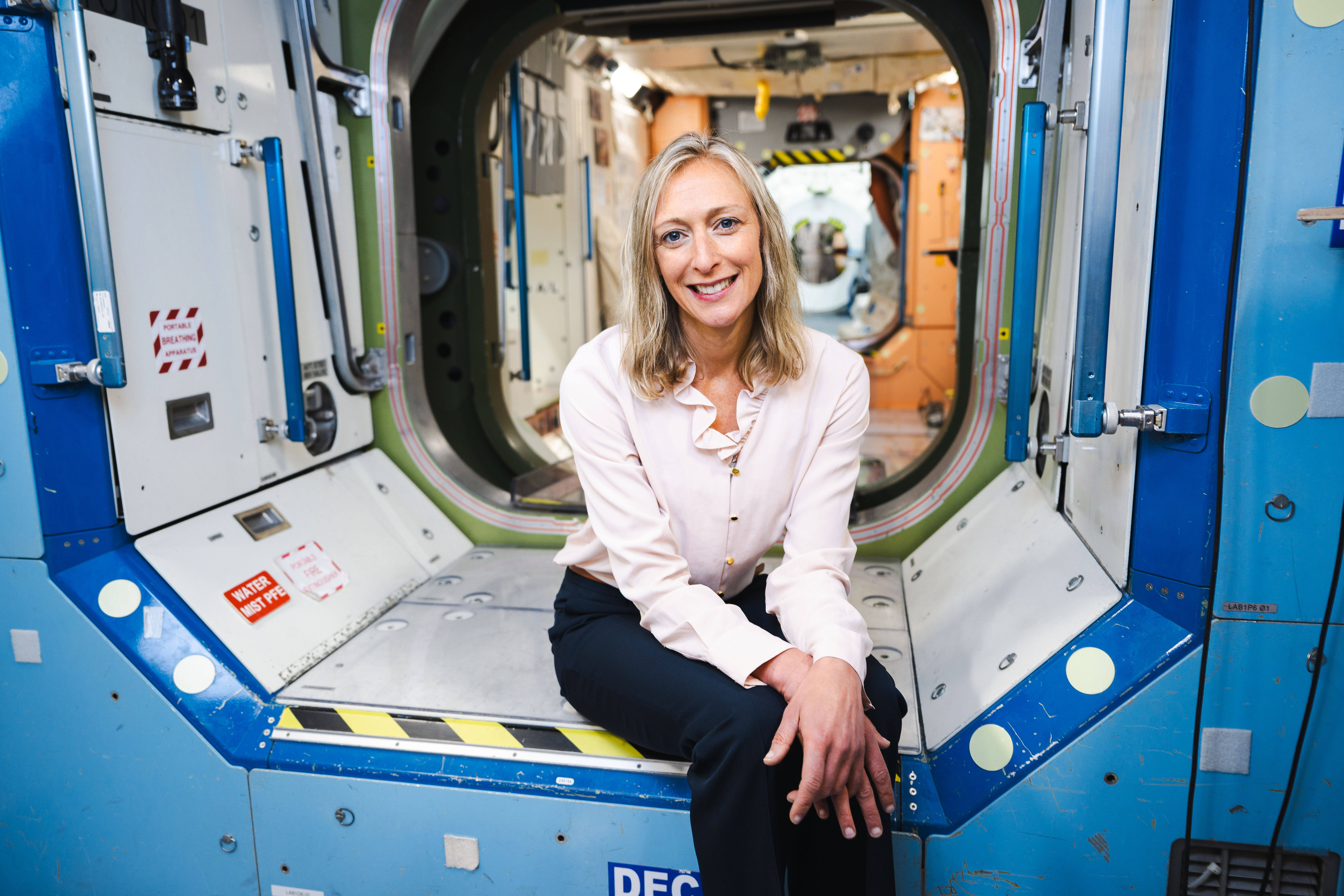 Meghan Everett, wearing dark dress pants and a light-colored blouse with buttons and a ruffled collar, smiles at the camera as she sits at the edge of one of the International Space Station modules inside the Space Vehicle Mockup Facility at NASA's Johnson Space Center. The edges of the module mockup are blue, with a yellow and black striped warning strip present on the step where Meghan is sitting. Blue handrails are present on both sides of the module mockup, and a wide variety of labels are visible on all of the module mockup surfaces within.