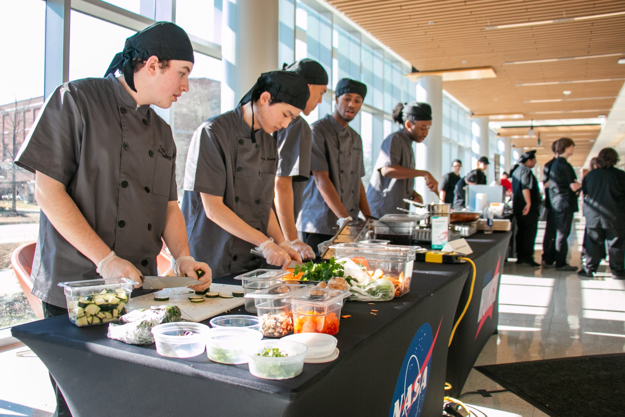 High school students in matching grey aprons and black chefs hats chop vegetables on a long black table as they prepare their savory entry for NASA's HUNCH Culinary Challenge