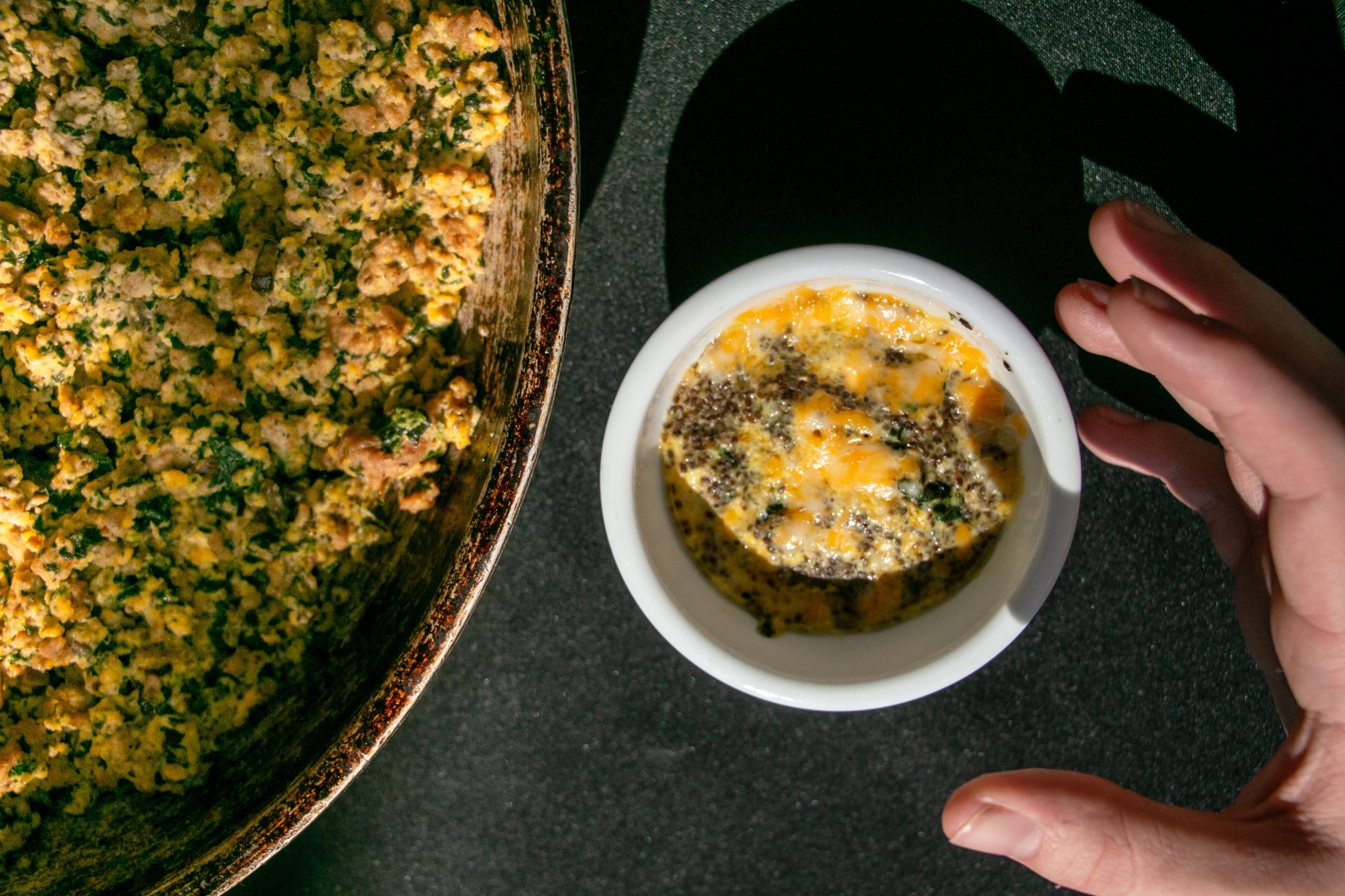 An eager hand reaches for a small serving of eggs scrambled with vegetables and topped with seeds as a larger skillet of the savory breakfast dish sits to the left.