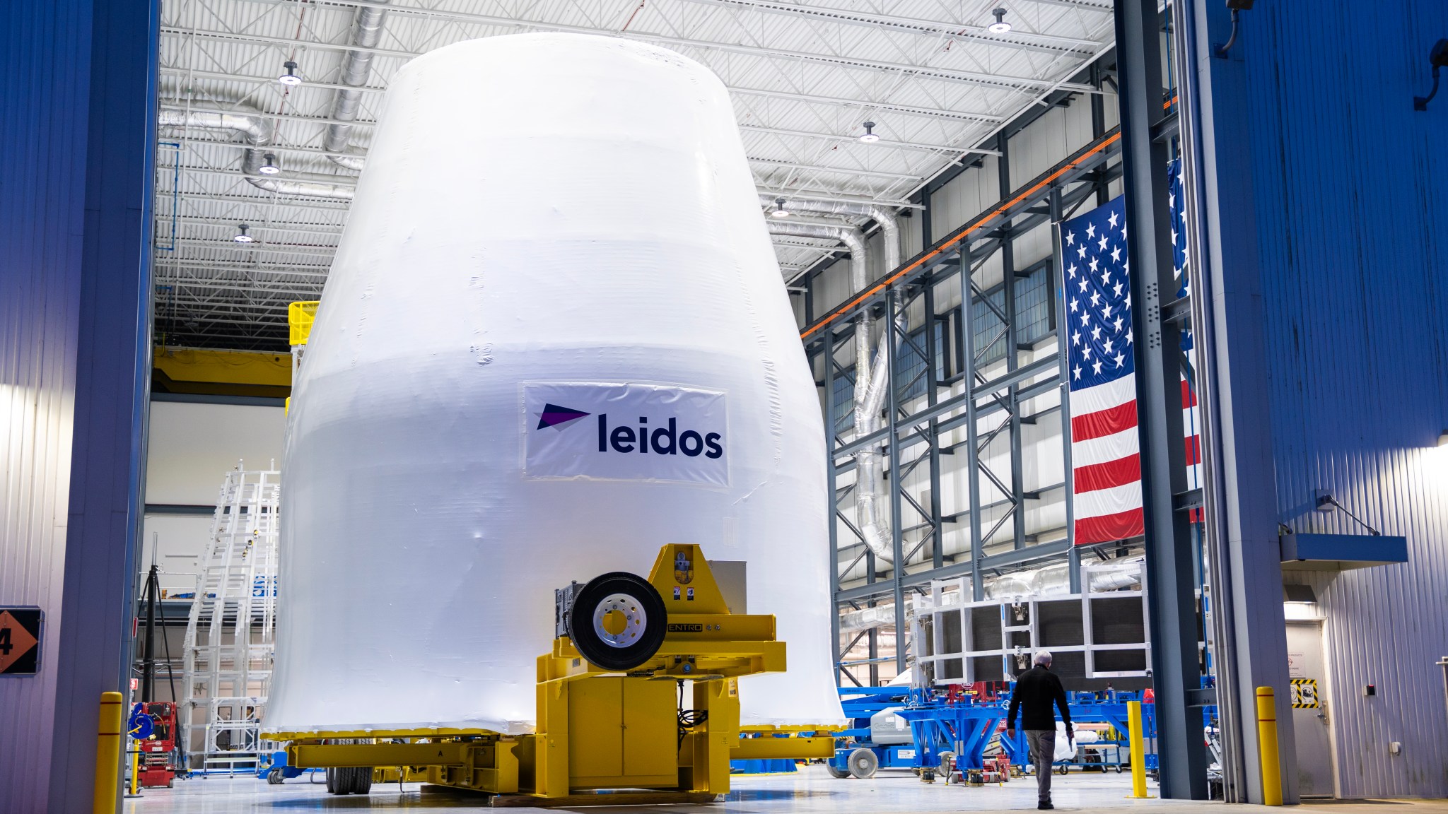A test version of the universal stage adapter for the SLS (Space Launch System) rocket for Artemis 4 is seen inside Marshall Space Flight Center’s facility in Huntsville, Alabama. The adapter sits on a yellow piece of hardware. There is an American flag hanging on the wall to the right and the word “Leidos” is painted black on the white adapter.