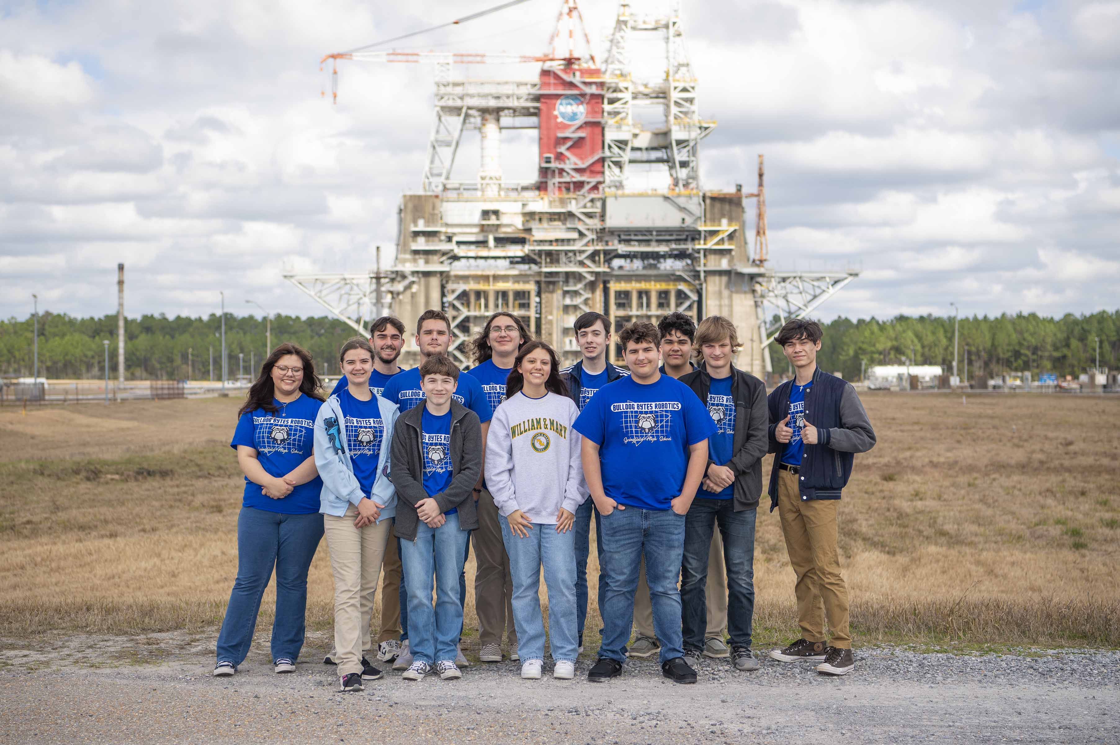 two rows of high school students wearing blue t-shirts stand in the forefront with the Thad Cochran Test Stand in the background