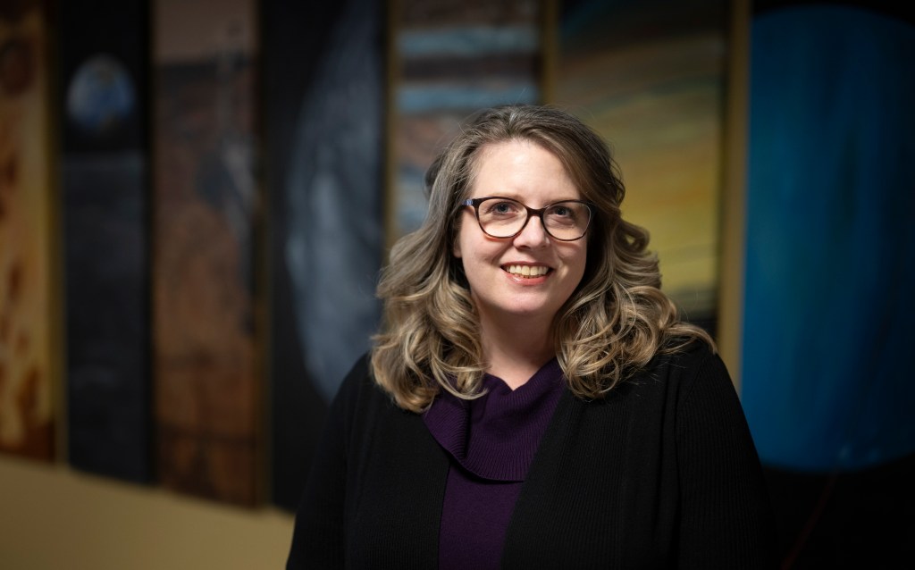 A woman wearing thick-rimmed glasses, a purple sweater, and a black cardigan smiles widely at the camera. Paintings of worlds across our Solar System are blurred in the background as light from above perfectly frames her face, surrounded by loose, dirty blonde curls.