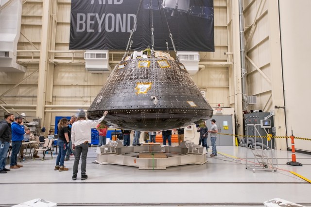 Inside the high-bay assembly area in the Space Environments Complex at NASA’s Neil Armstrong Test Facility, the charred Orion capsule from Artemis I is hoisted about four feet above the round white metal framing that it will be tested in. Several engineers and technicians wearing jeans, casual shirts, and work boots surround the capsule.