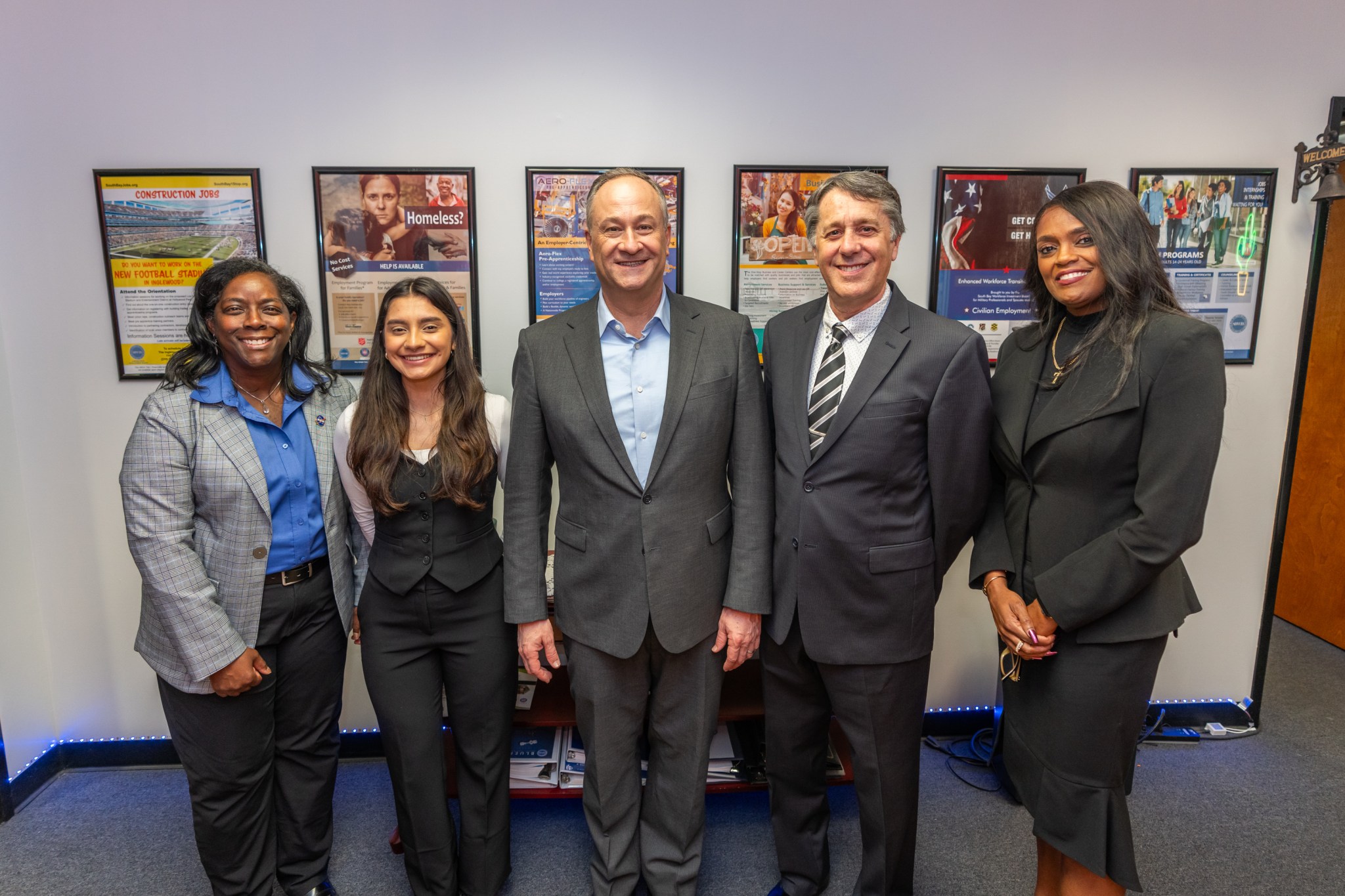 Three women and two men stand in a row. The white wall behind them has six posters of various subjects hung in a row.