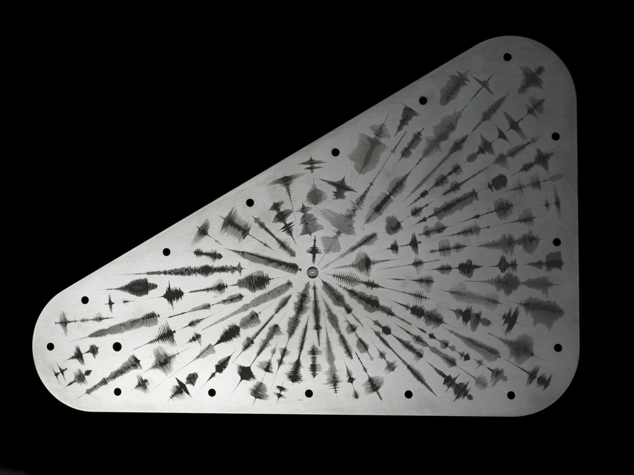 A triangle-shaped metal plate with rounded edges against a black background. The silver-colored plate has waveforms, visual representation of sound waves, radiating out from a circular symbol toward the edges of the plate.