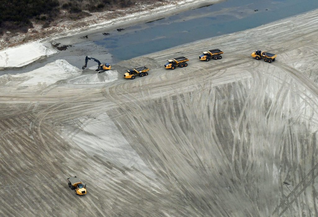 One full dump truck, backhoe excavating sand from Wallops beach and loading sand into a dump truck, three empty dump trucks waiting to be filled with sand.