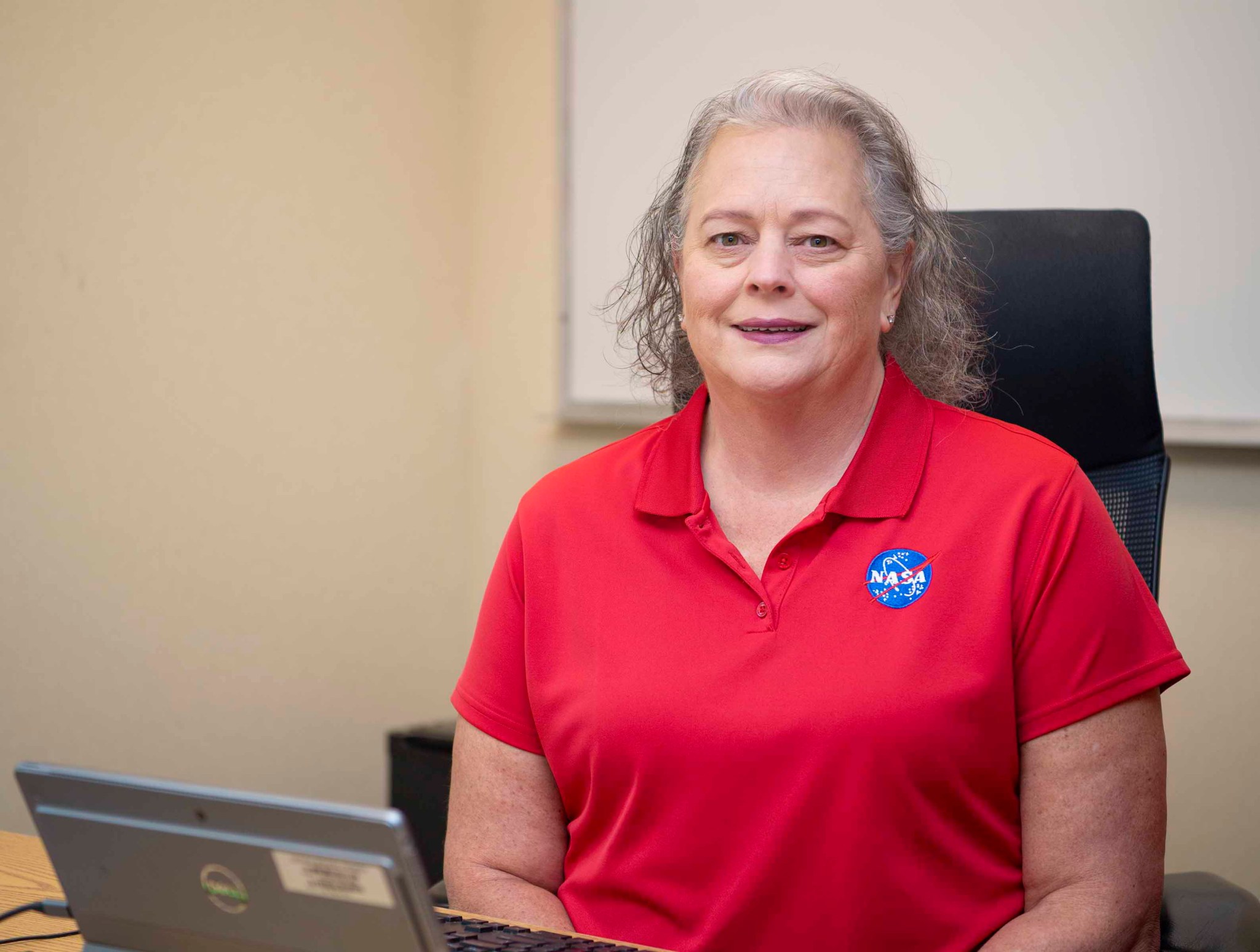 Rae Anderson, wearing a red polo shirt with the NASA meatball on the pocket, smiles at the camera while using her laptop.