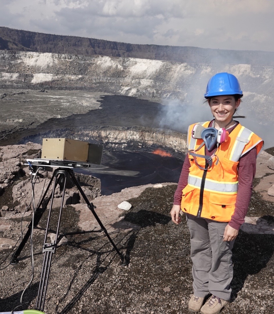 Casey Honniball wears a blue hard hat and orange vest while standing in front of a field work site. 