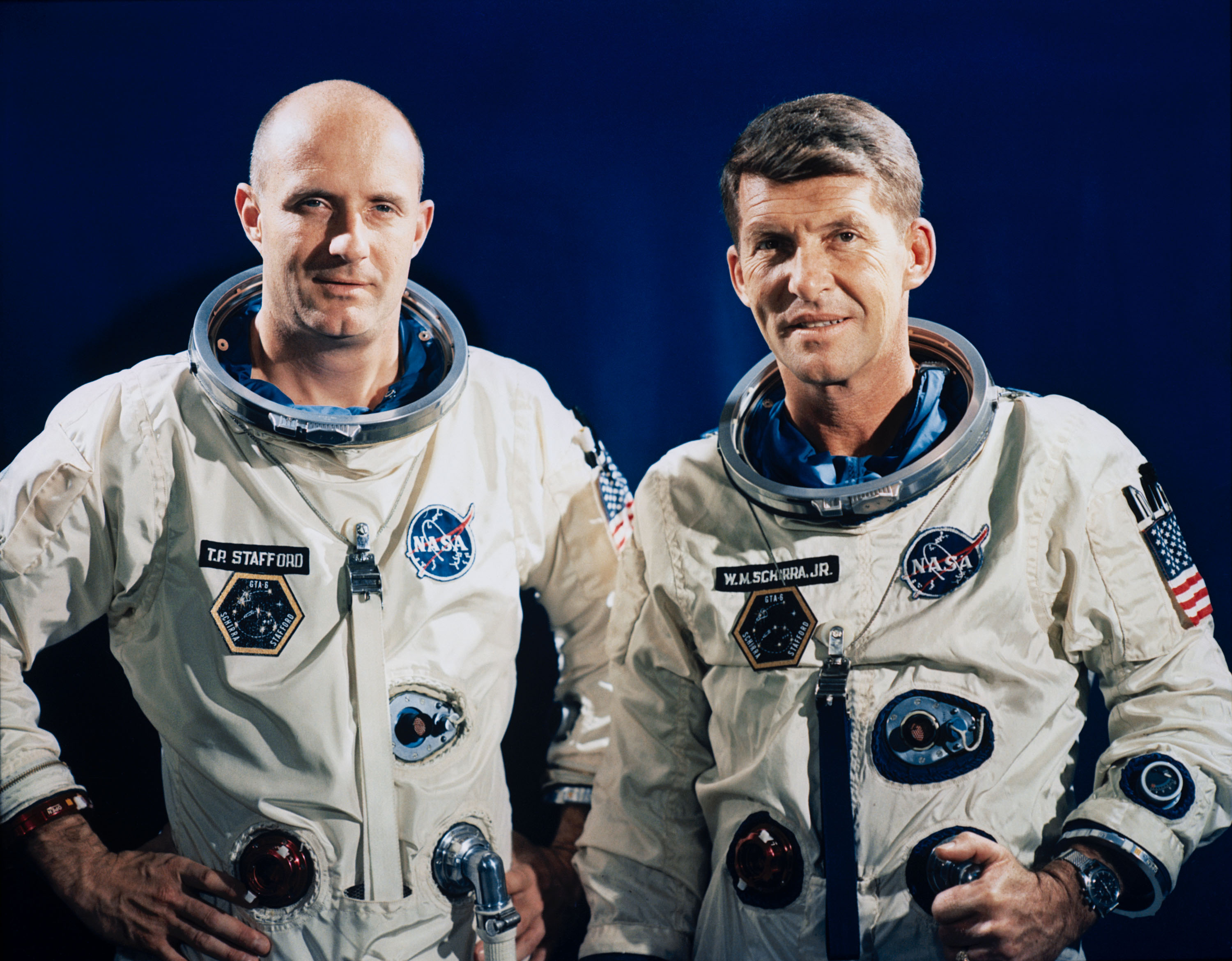 Two male astronauts, Thomas P. Stafford (left) and Walter M. Schirra Jr., look directly into the camera. They are wearing white spacesuits with multiple patches including their names, mission, NASA, and the American flag. Behind them is a deep blue backdrop.