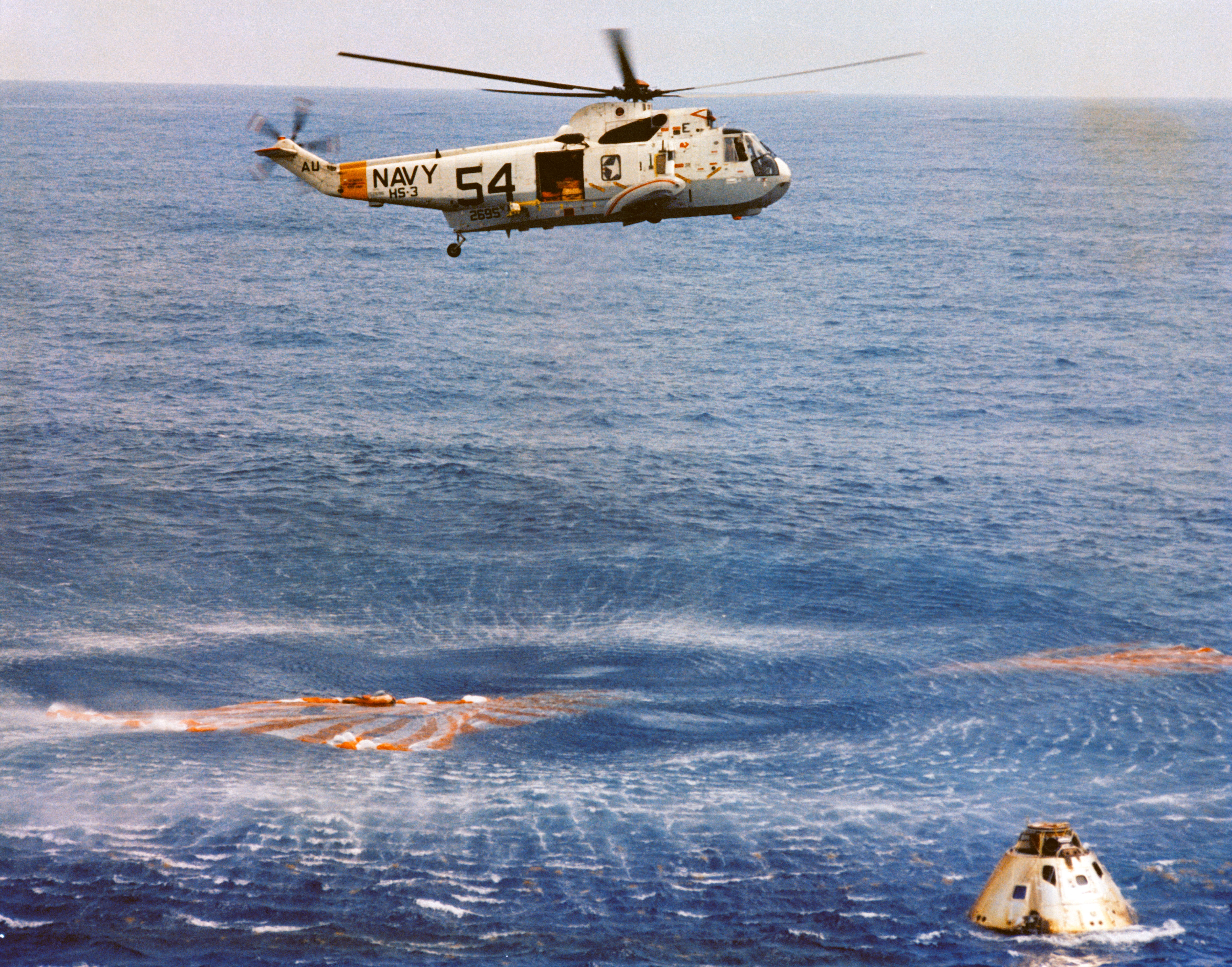 A recovery helicopter with "Navy" and "54" stenciled on it hovers above the water, the wind from its blades creating rings of circles below. Directly below the helicopter is an orange and white parachute. At bottom right is the Apollo 9 command module, where the astronauts await recovery.