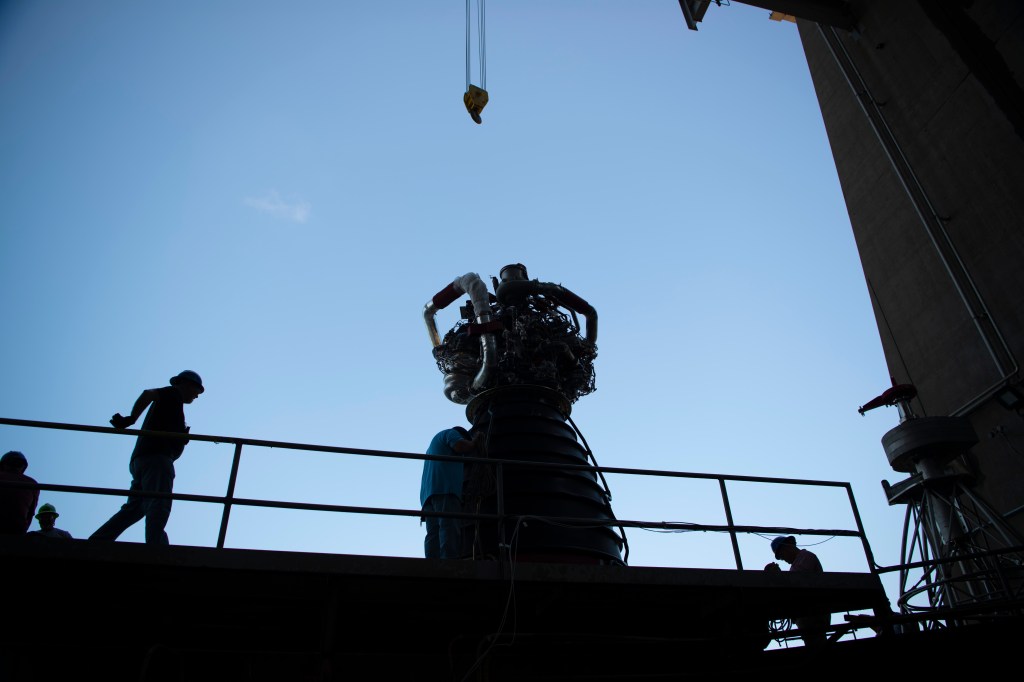 image is the silhouette of a RS-25 and two workers on the test stand