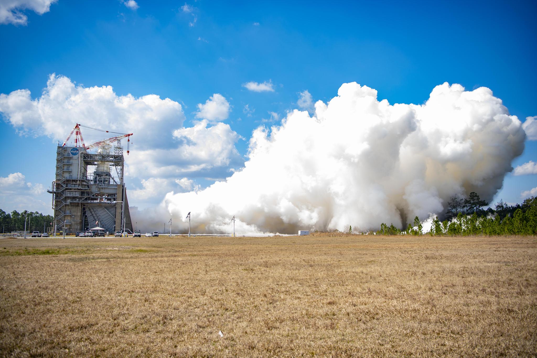 vapor clouds erupt from a RS-25 hot fire; view is across a grassy field