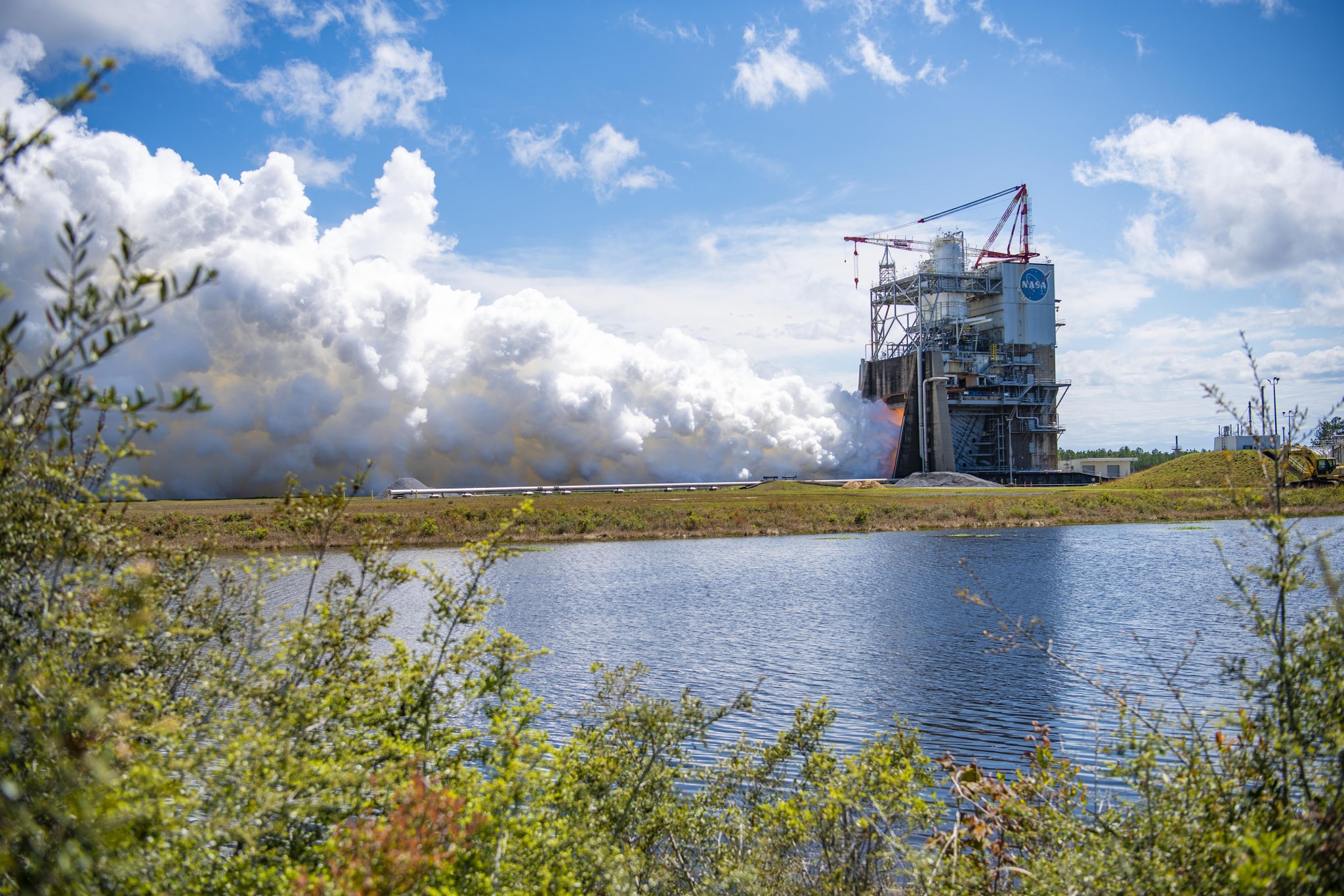 a view of the Fred Haise Test Stand during a hot fire in the background framed by green brush and young trees in the forefront.