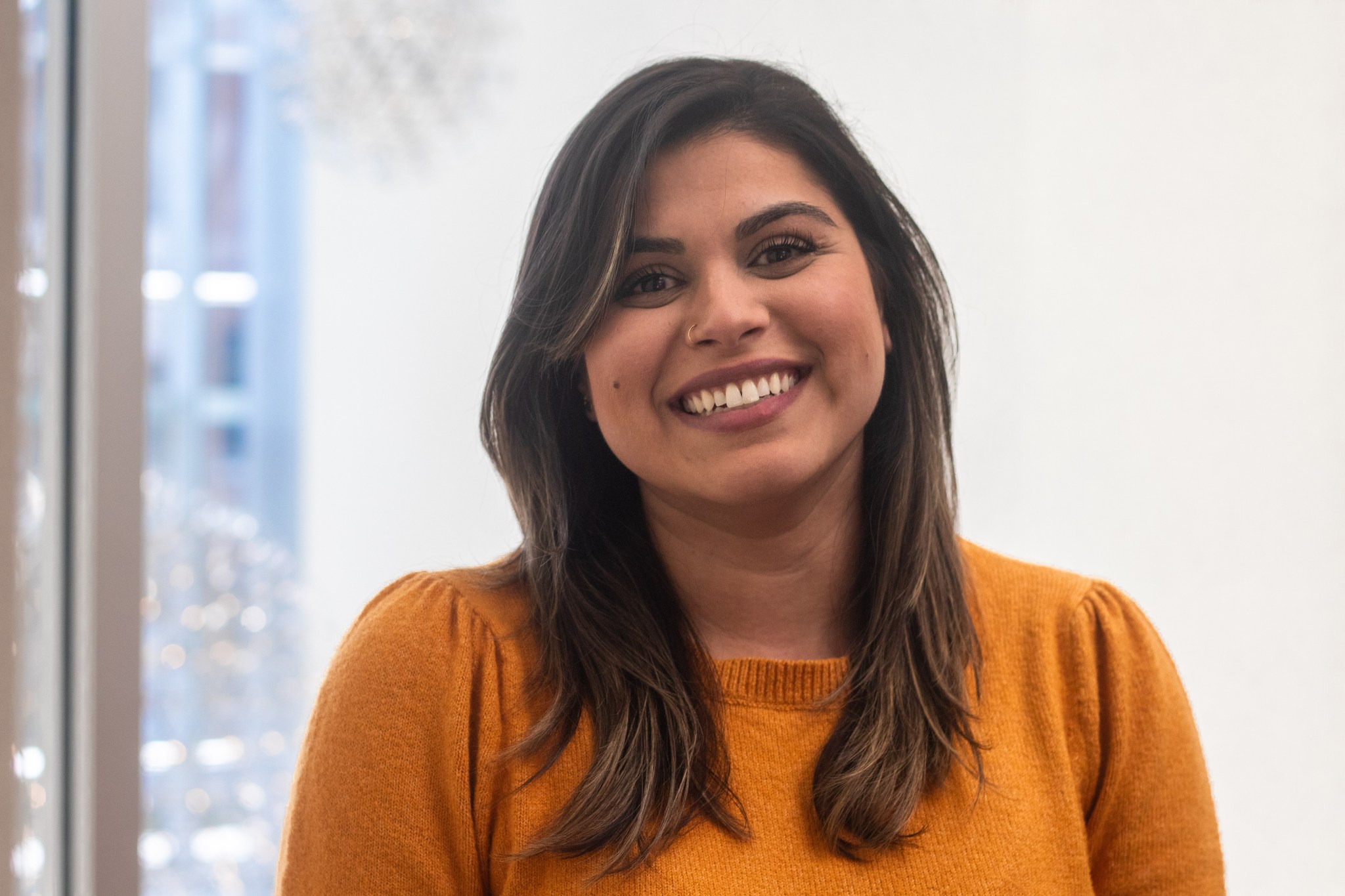 Anum Ashraf is a Climate Scientist at NASA Langley Research Center. She is pictured here smiling and wearing a mustard yellow sweater. Large windows can be seen behind her.