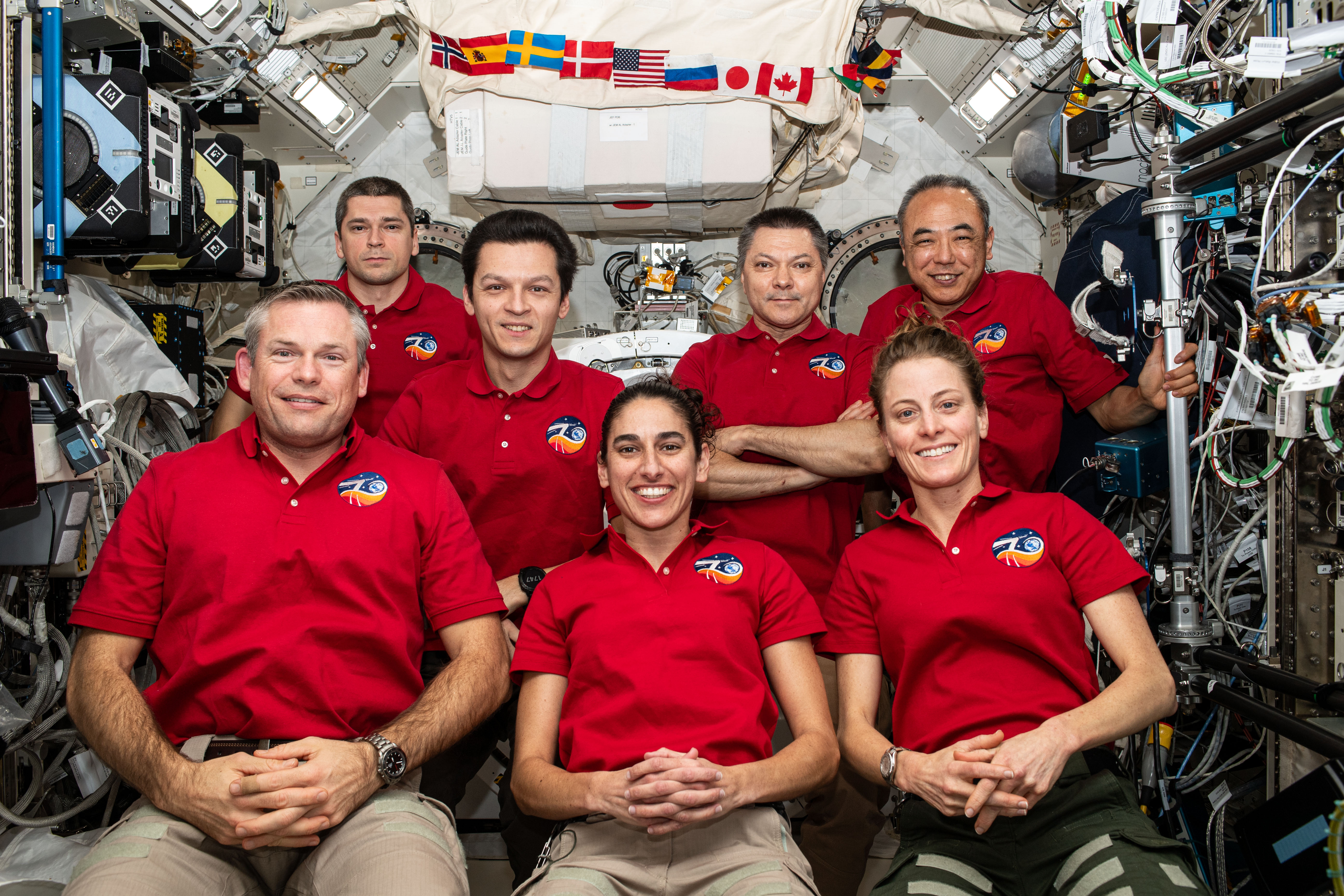 NASA astronauts Jasmin Moghbeli, front row center, and Loral A. O’Hara, front row right, and their Expedition 70 crew mates chat with space station program managers to celebrate the 25th anniversary of the orbiting laboratory.