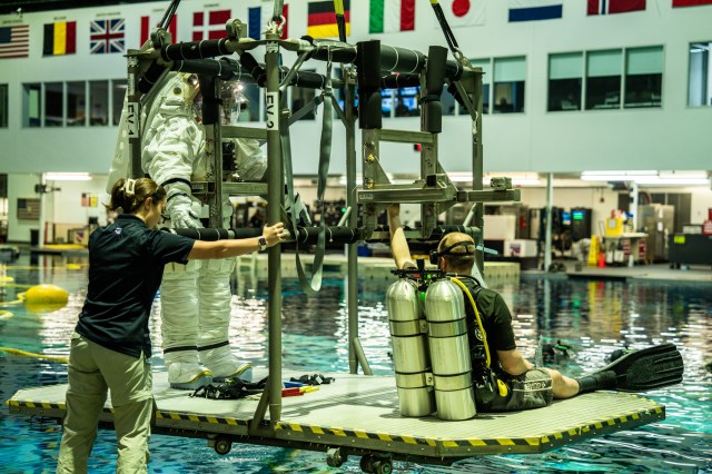 Axiom Space's AxEMU (Axiom Extravehicular Mobility Unit) spacesuit being tested at NASA Johnson's Neutral Buoyancy Laboratory. The white spacesuit is on a donning stand with a scuba diver about to be submerged underwater.