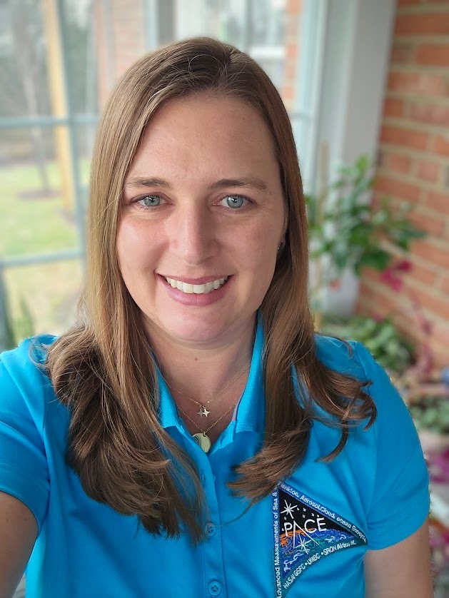 Kate A. McGinnis smiles for a selfie in a room with a brick wall, in front of a window and plant. She's wearing a blue polo with the PACE mission patch.