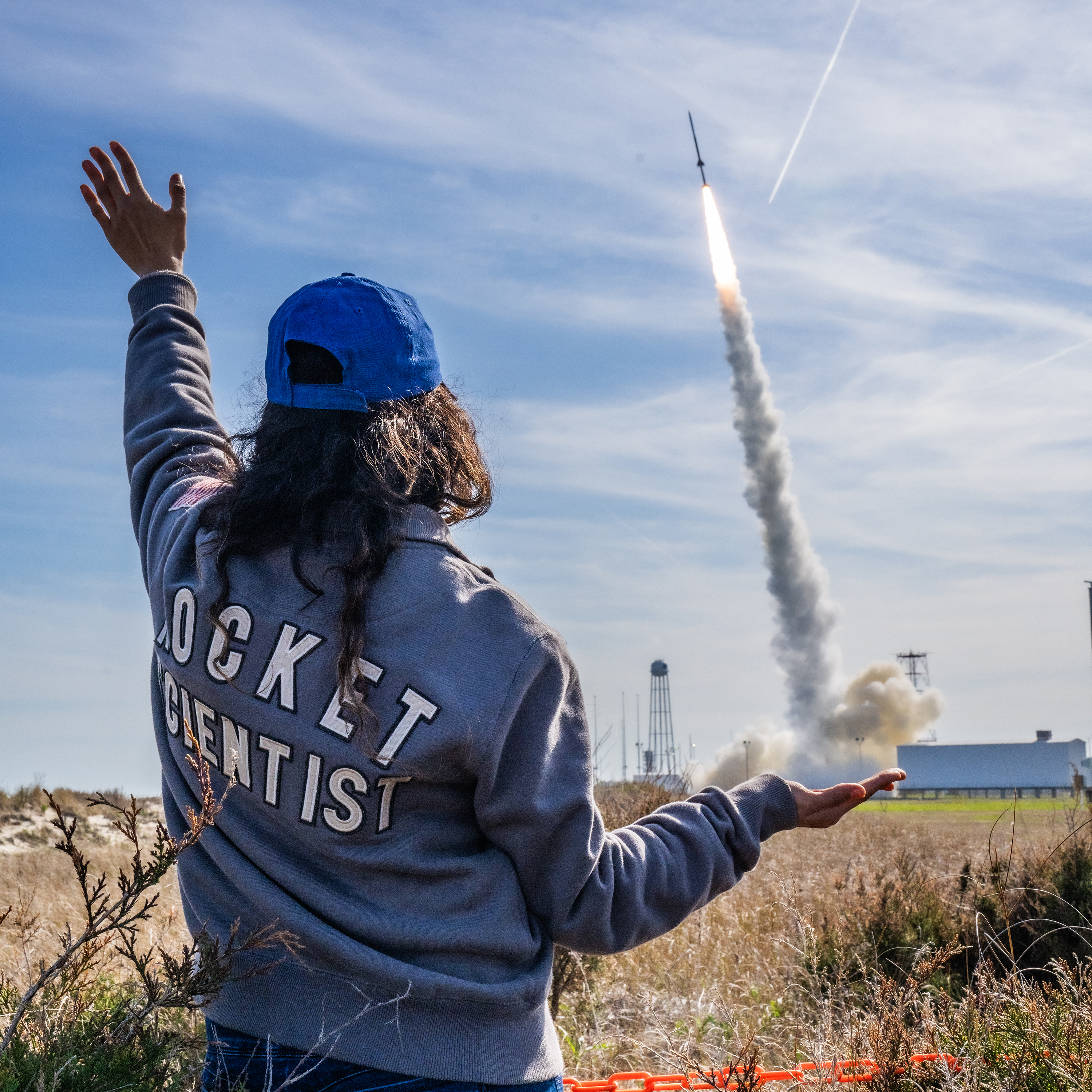 Cindy Fuentes Rosal, wearing a light-blue jacket with the words "Rocket Scientist" in white on the back, faces away from the camera with her hand in the air. In the background is a sounding rocket launching from a launch pad with a thick white trail of smoke underneath.