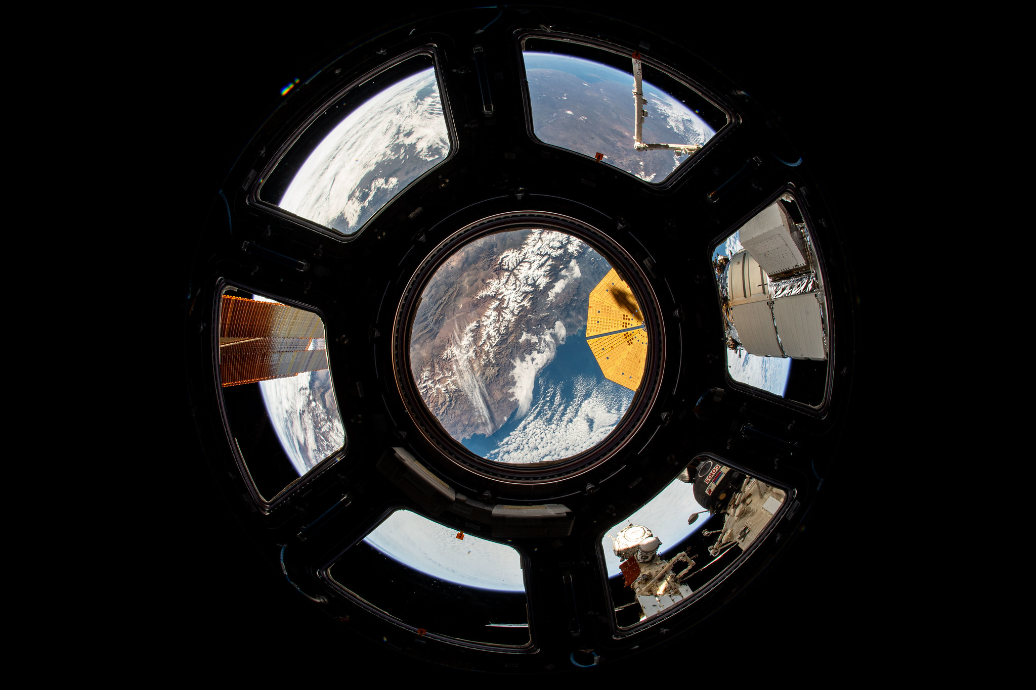 Peering through the International Space Station's Cupola, or "window to the world," the western coast of Chile is visible. Through a window on the left, one of the orbiting laboratory's solar arrays can be seen; in the center, Northrop Grumman's cymbal-shaped solar array is peeking in, and in the bottom right corner window, the Roscosmos segment of the station is visible.