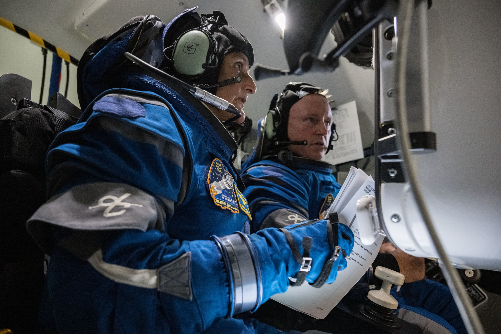 Williams, seated in the foreground, and Wilmore, seated next to her, wear blue spacesuits, gloves, and headsets as they study a monitor in front of them. Williams is holding a sheaf of papers in her right hand.