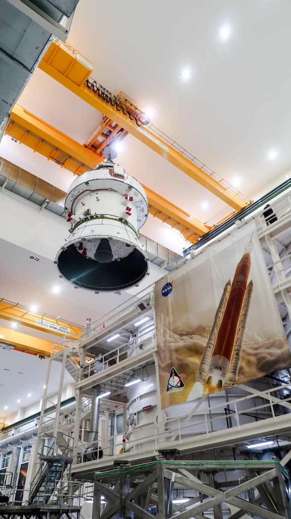 An Orion spacecraft is suspended by a crane mounted inside a building and hoisted for stacking atop another spacecraft component.