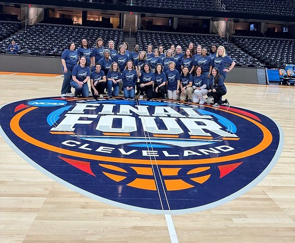 Three rows of women in blue shirts with NASA emblems pose for a photo on center court inside Rocket Mortgage FieldHouse. The large Final Four emblem is on the floor in front of them and empty stands can be seen behind them.