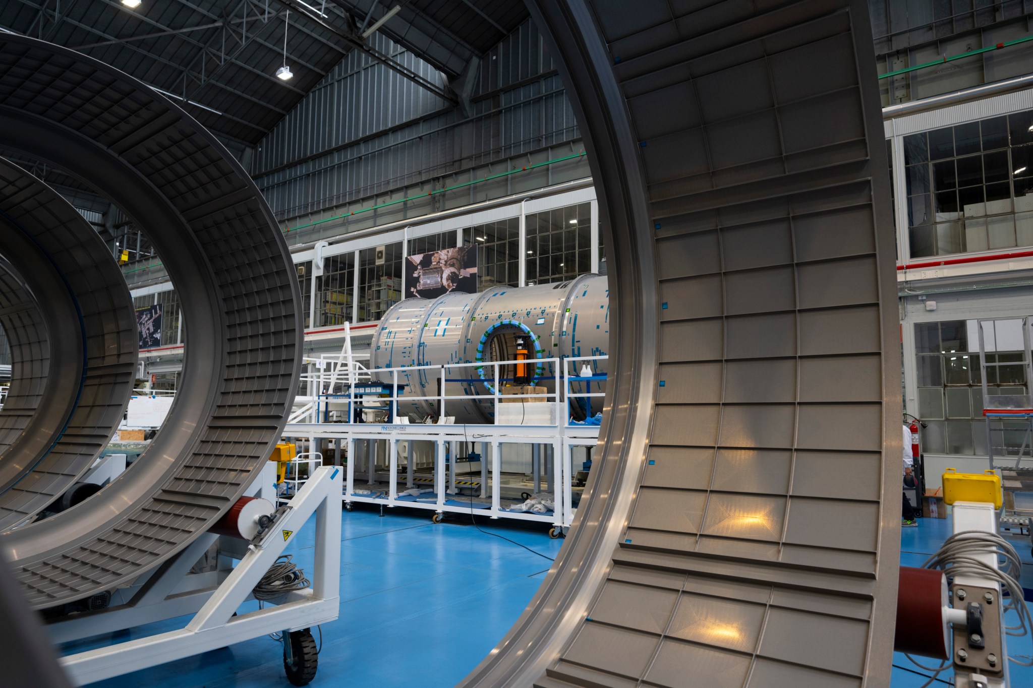 In the background, a large space station module for Gateway in a facility ringed by inspection scaffolding. In the foreground, large gray rings.