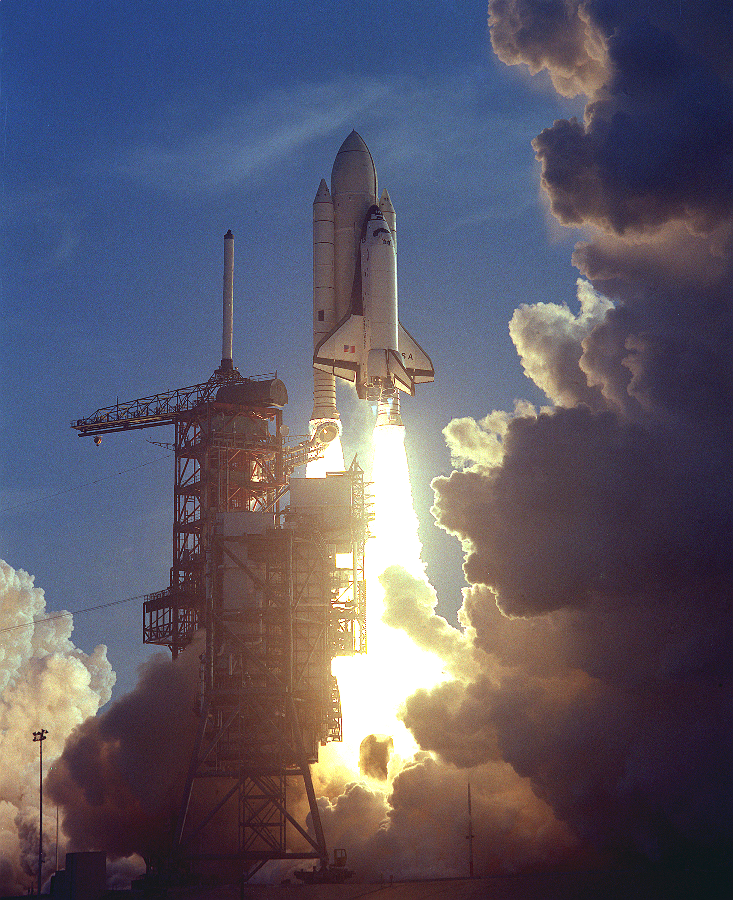 The first space shuttle takes off from a launch pad. White clouds billow upward, obscuring the rightmost third of the image. Bright flames pour out from the rockets; in contrast, the rest of the image is dim.