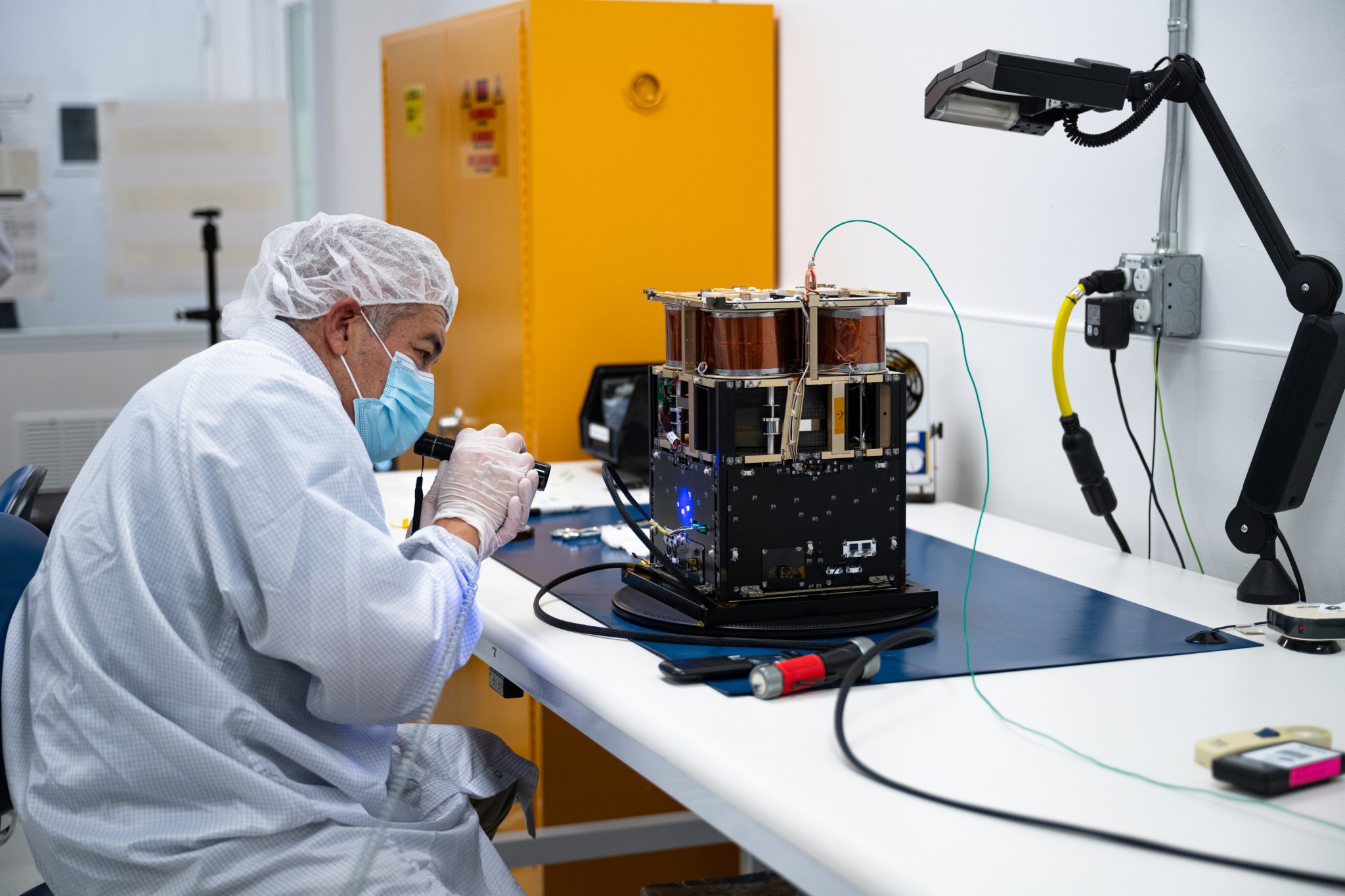 A man in protective gear, including a hair net and face mask, inspects the Advanced Composite Solar Sail System spacecraft with a UV flashlight in a laboratory environment.