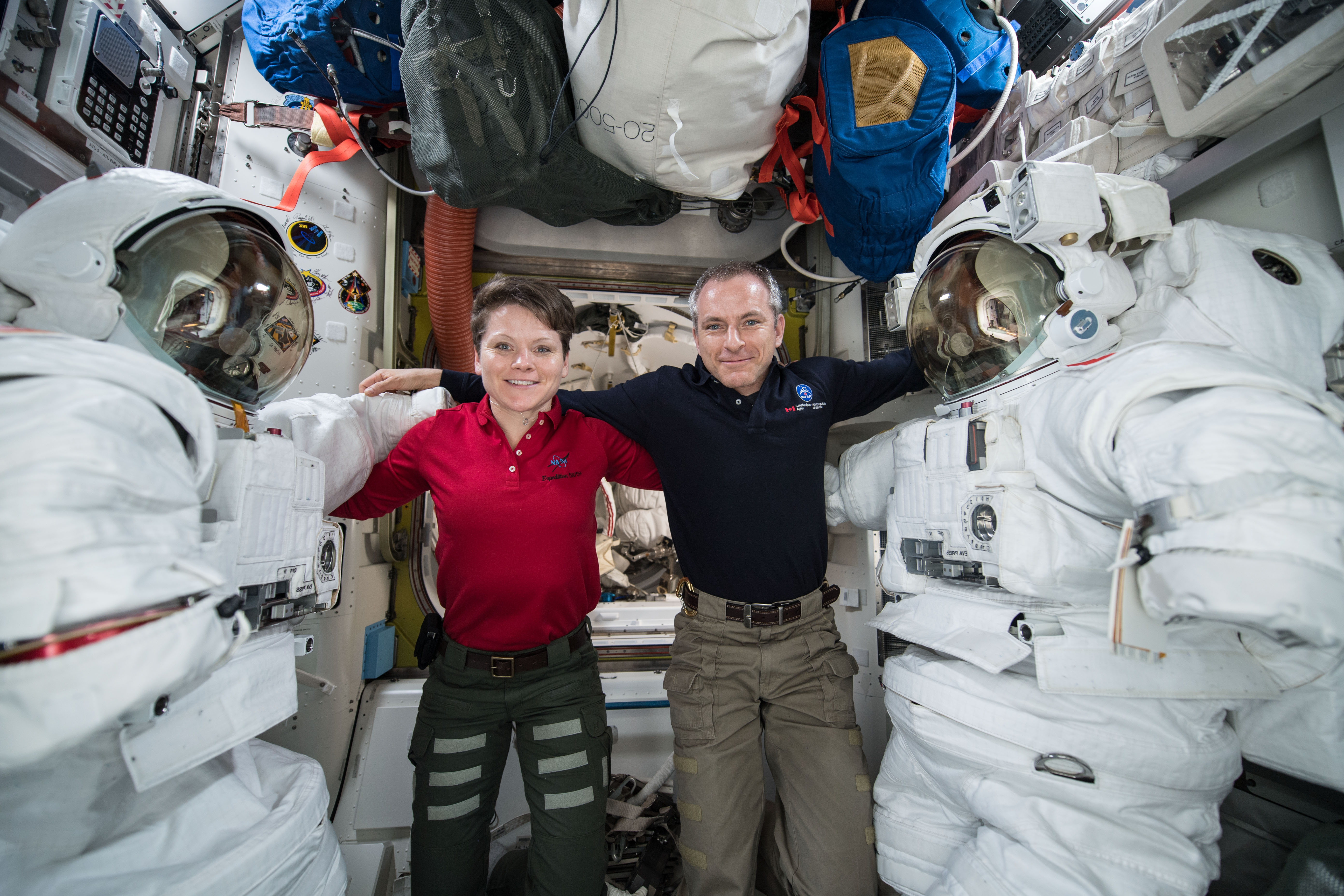 iss058e013453 (Feb. 14 2019) --- Astronauts (from left) Anne McClain and David Saint Jacques are pictured in between a pair of spacesuits that are stowed and serviced inside the Quest airlock where U.S. spacewalks are staged.