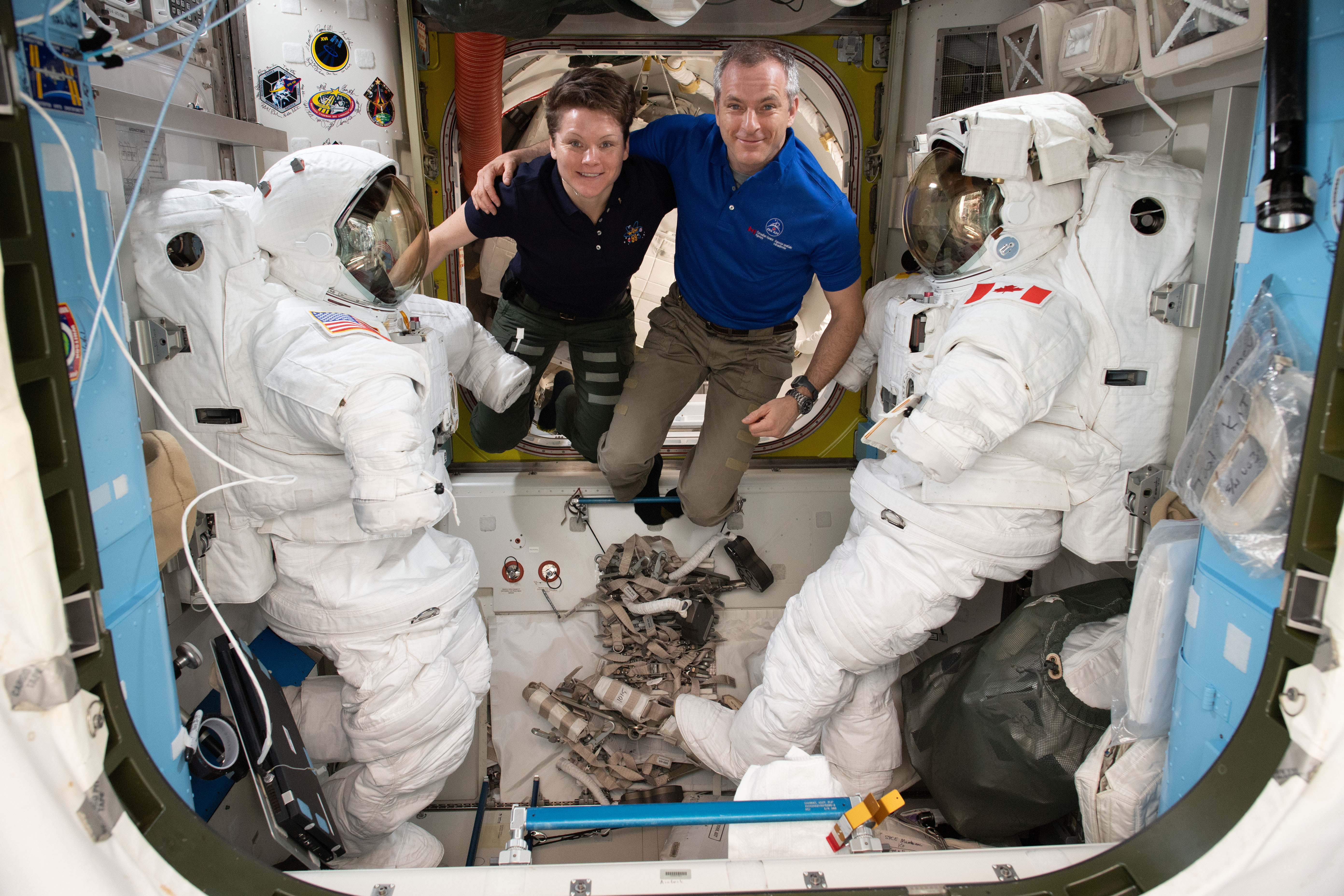iss058e015358 (Feb. 17 2019) --- Astronauts (from left) Anne McClain and David Saint Jacques are pictured in between a pair of spacesuits that are stowed and serviced inside the Quest airlock where U.S. spacewalks are staged.