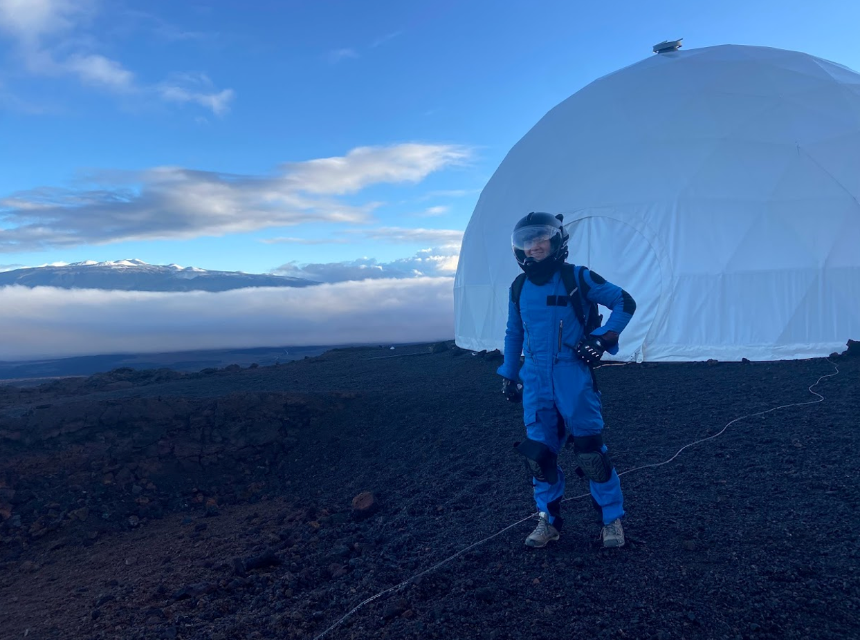 Katie Fisher stands in front of the HI-SEAS habitat in an EVA suit with Mauna Kea in the background.