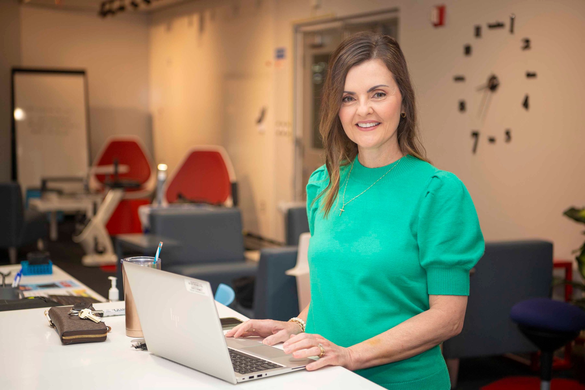 a woman, cherie beech, wearing a green sweater smiles at the camera while standing at a desk working on a laptop