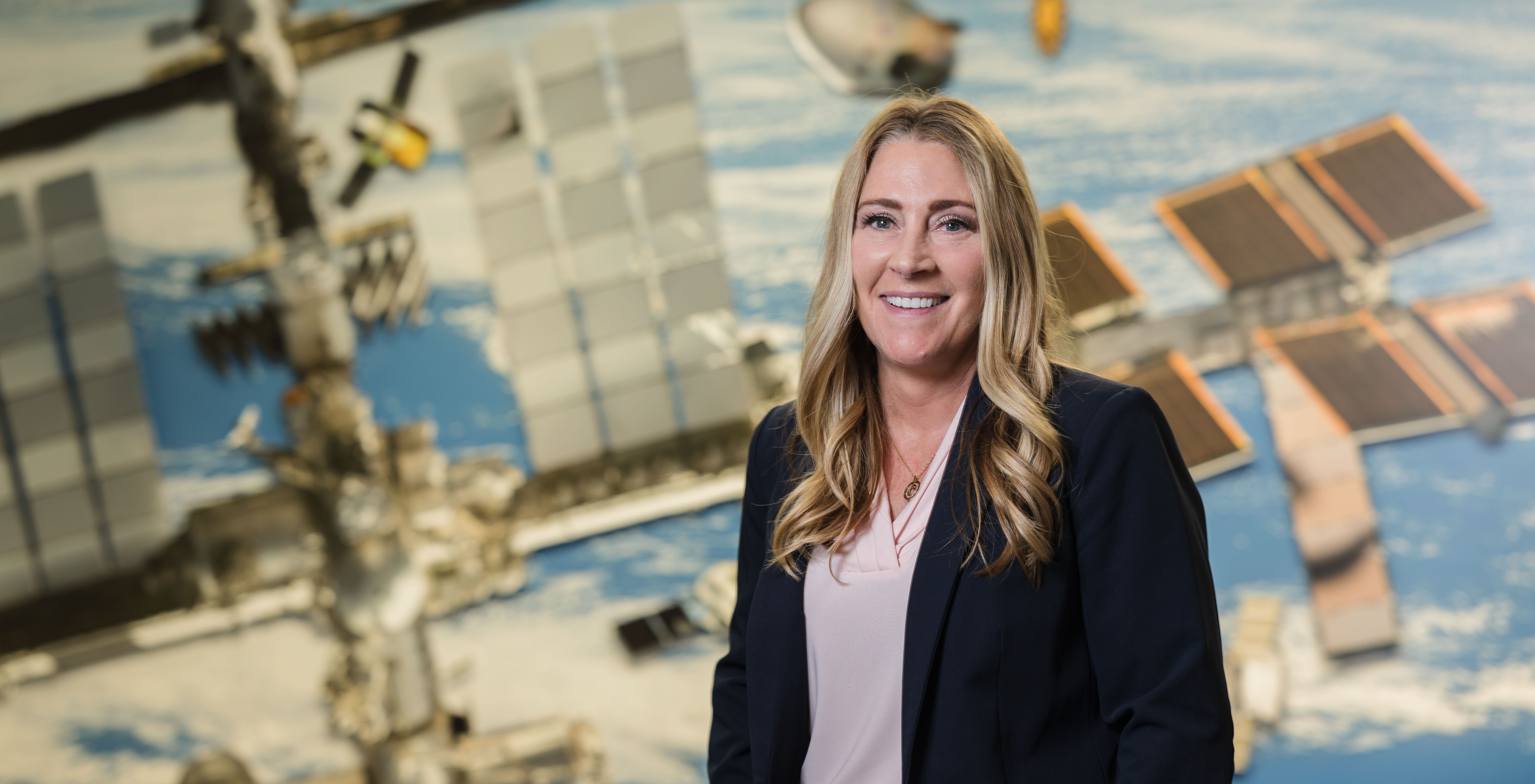 Dana Weigel stands and smiles in front of a large wall mural of the International Space Station. She has wavy blond hair and is wearing a dark blue jacket over a pink blouse with a circular necklace around her neck.