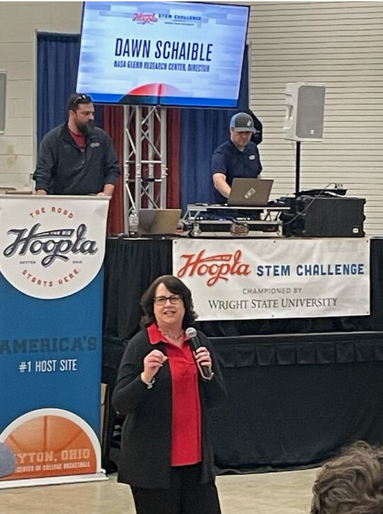 NASA Deputy Center Director Dawn Schaible talks into a microphone. Her name is illuminated on a screen above her, and Hoopla STEM Challenge banners can be seen behind her. Two men work sound and video logistics behind her.