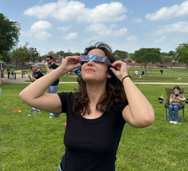 Emma Friedman, a NASA intern, is pictured here observing the total solar eclipse on April 8, 2024. Emma is standing on a grassy field wearing a black shirt and special eclipse sunglasses. A blue sky can be seen behind her.